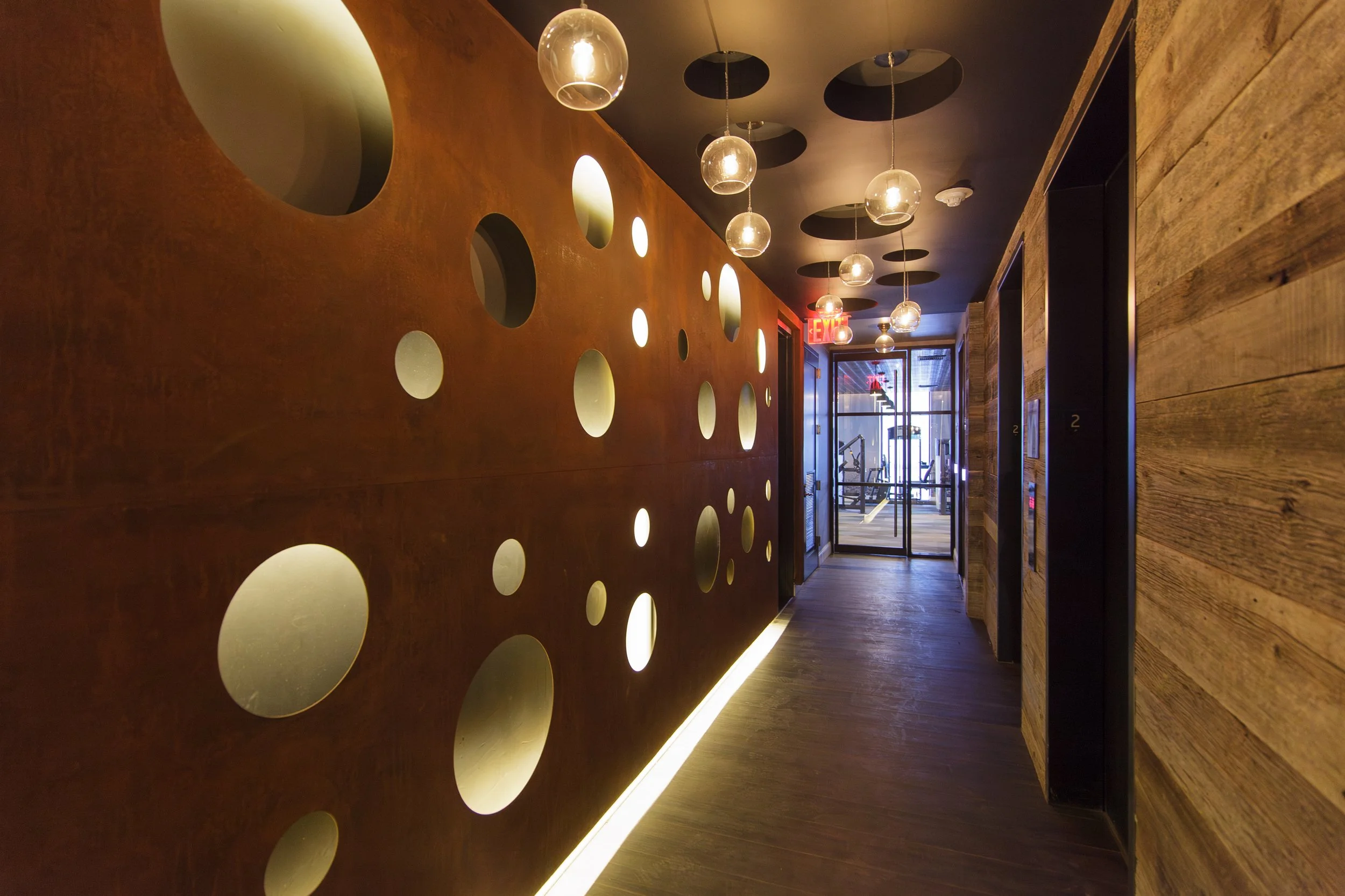 A modern hallway with a wooden wall featuring circular cutouts illuminated from behind, hanging spherical light fixtures, and a glass door at the end with an exit sign above.