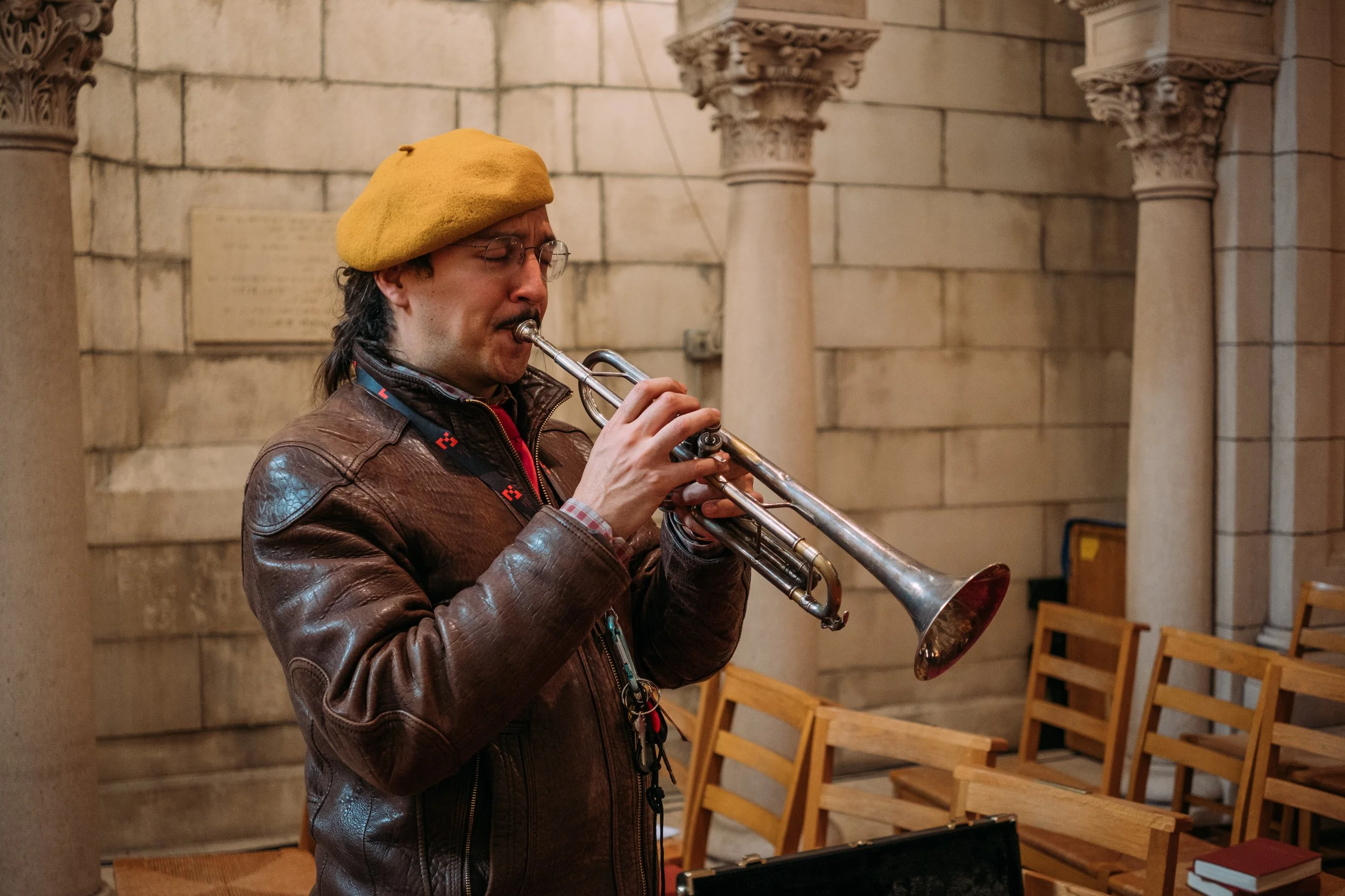 A man wearing glasses and a yellow hat playing a trumpet in an indoor setting with stone and brick walls and wooden chairs.