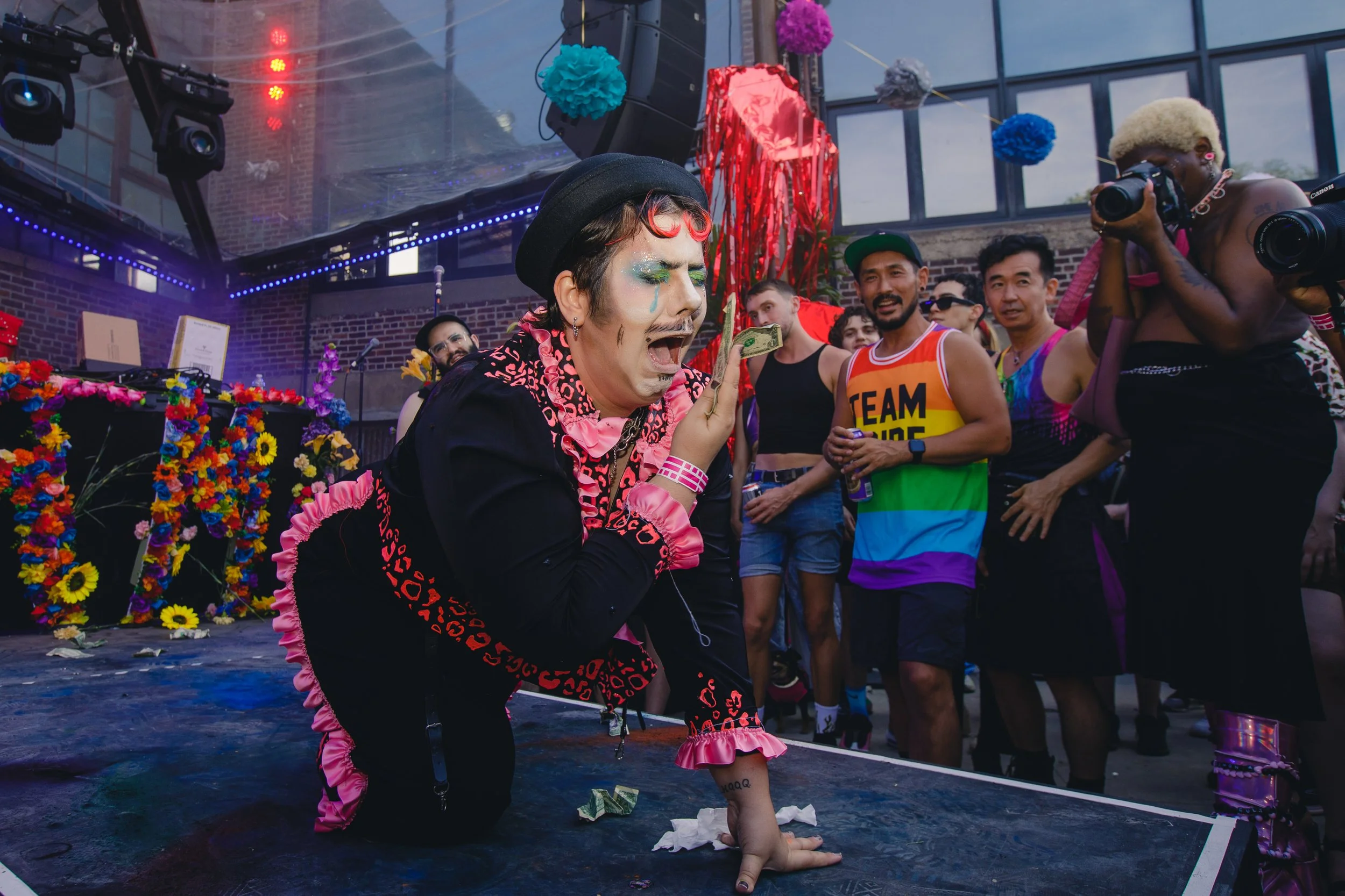 A person dressed in colorful, unusual clothing and makeup, crawling on the ground with a dollar bill on their face, surrounded by a crowd of diverse onlookers at an outdoor event with decorations, flowers, and stage lighting.