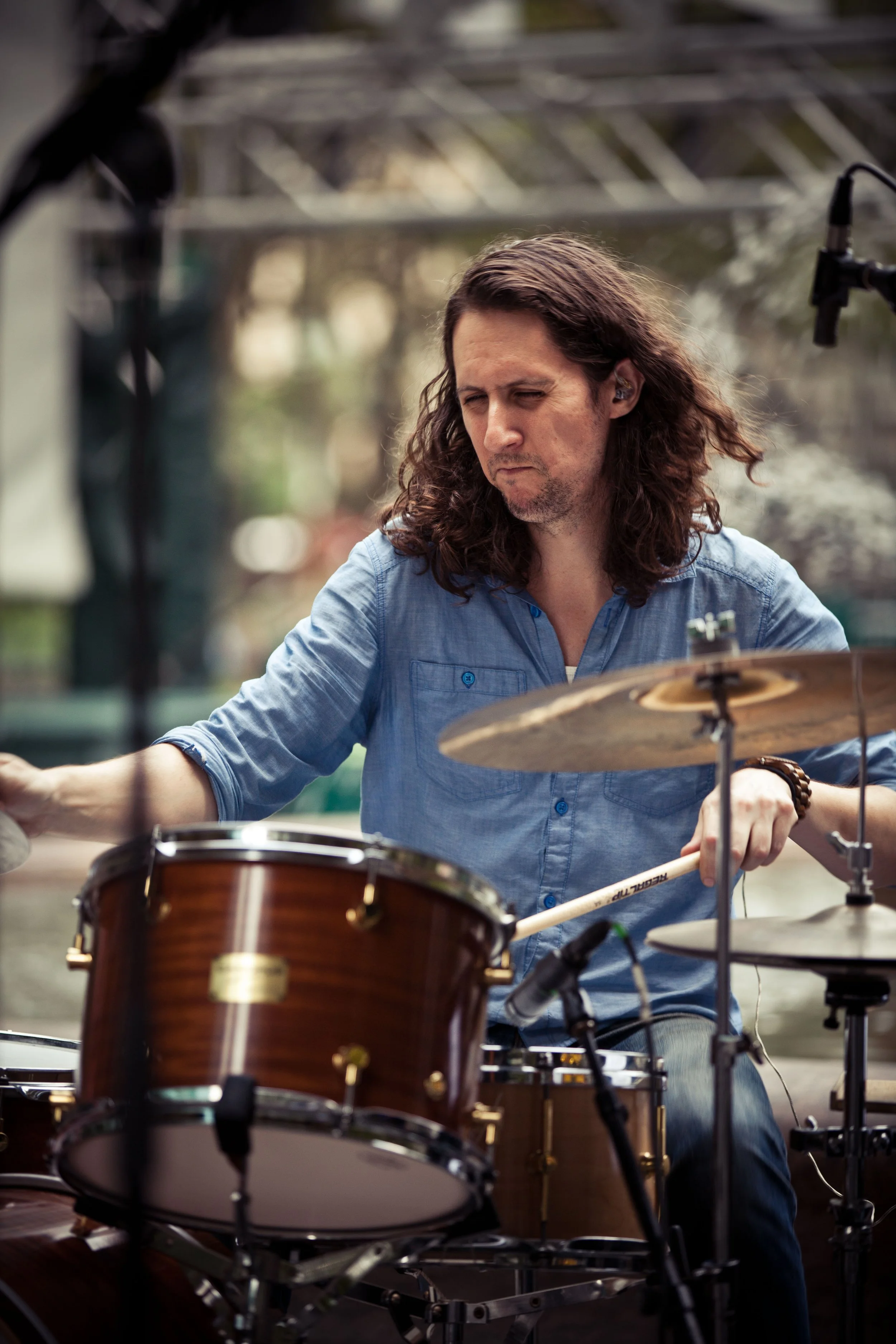 A man with long dark hair and a blue shirt playing drums outdoors. Focused expression, with drums and cymbals in front, microphone on microphone stand nearby, blurred background.