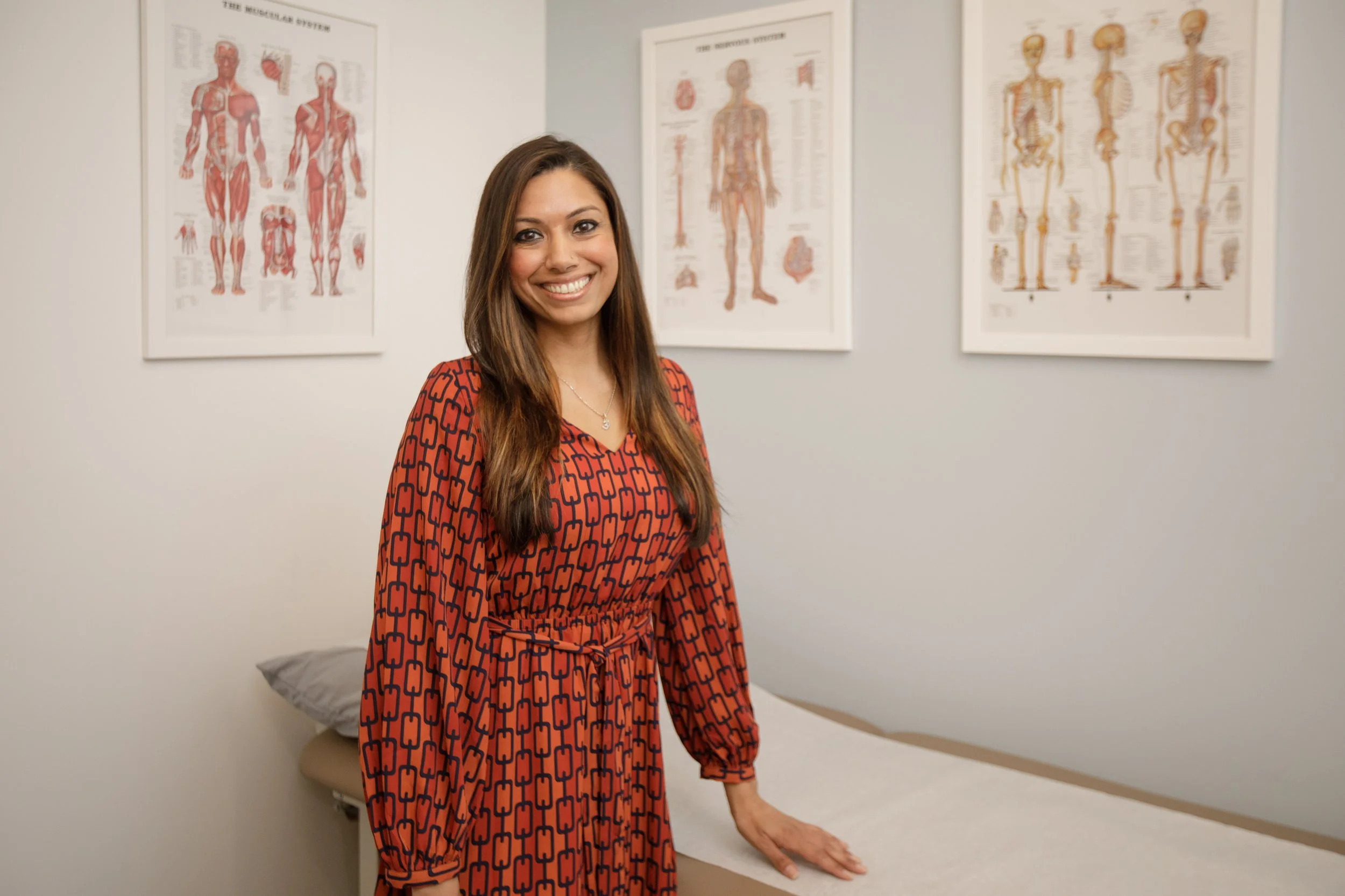 A woman with long brown hair smiling, wearing a red dress with a black geometric pattern, standing in a room with anatomical posters on the wall behind her.