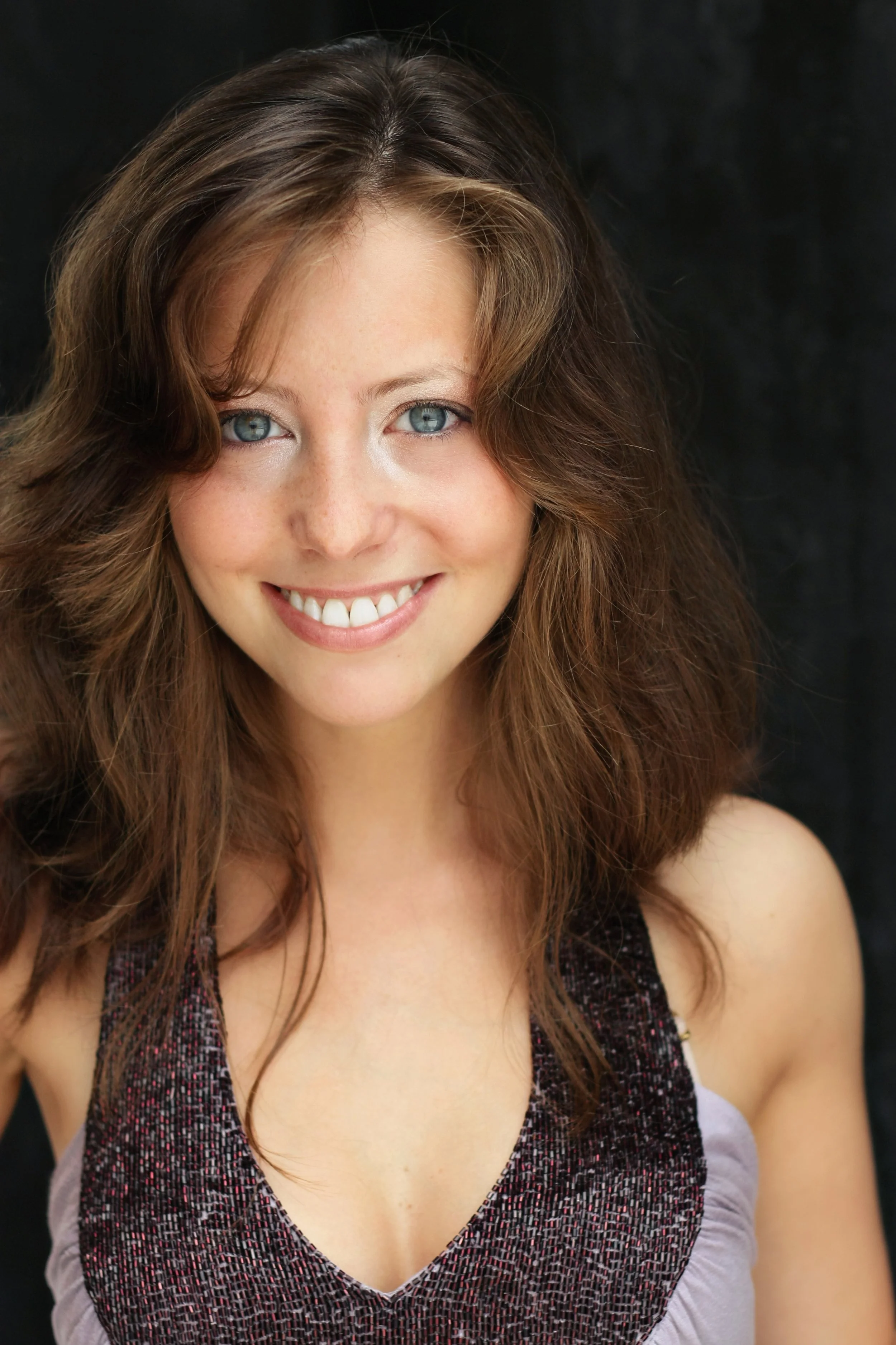 A smiling woman with blue eyes, long brown curly hair, wearing a sleeveless top with a textured pattern, against a black background.