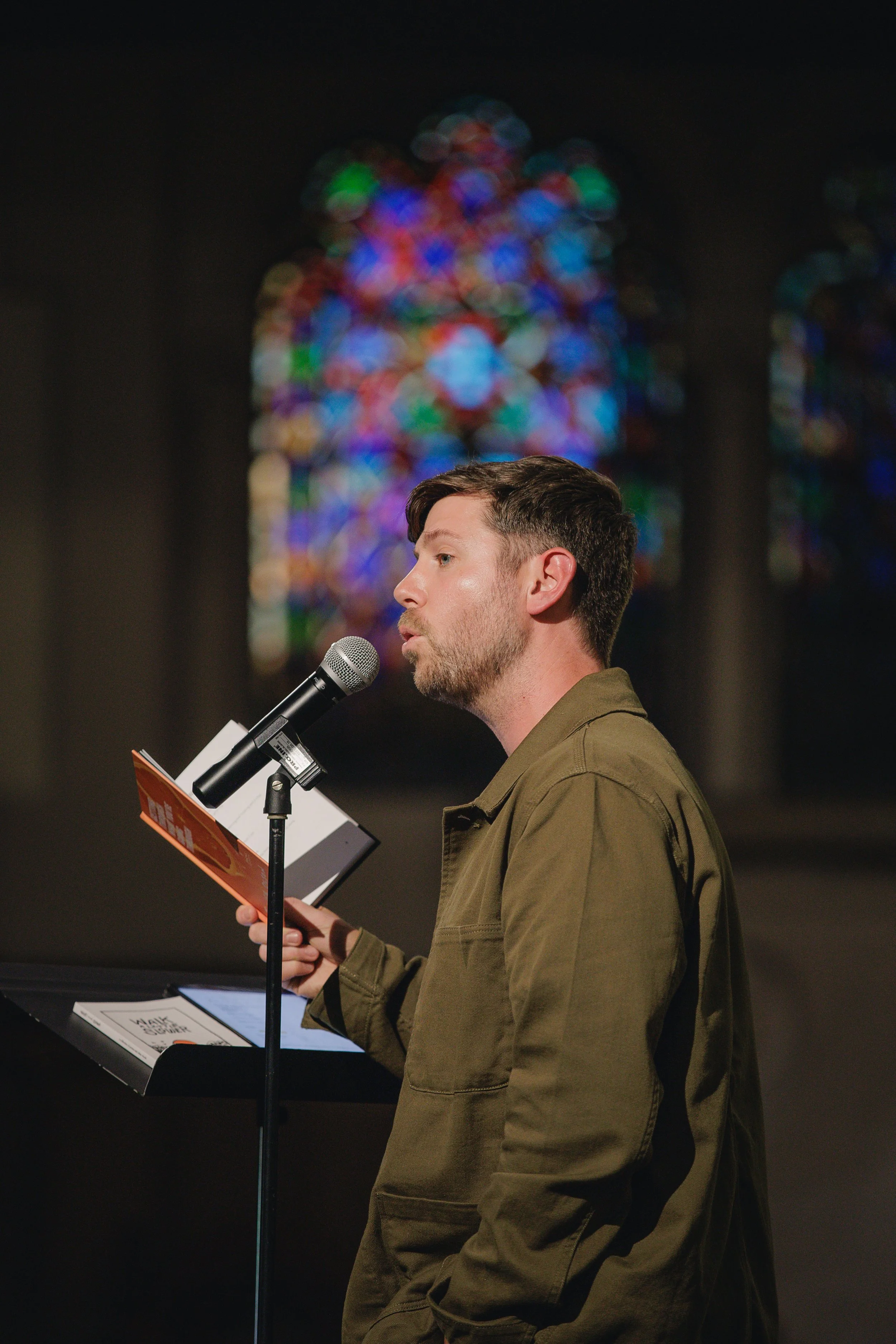 A young man with short dark hair and a beard, wearing an olive green jacket, speaking into a microphone while reading from a booklet or paper, with a colorful stained glass window in the background.