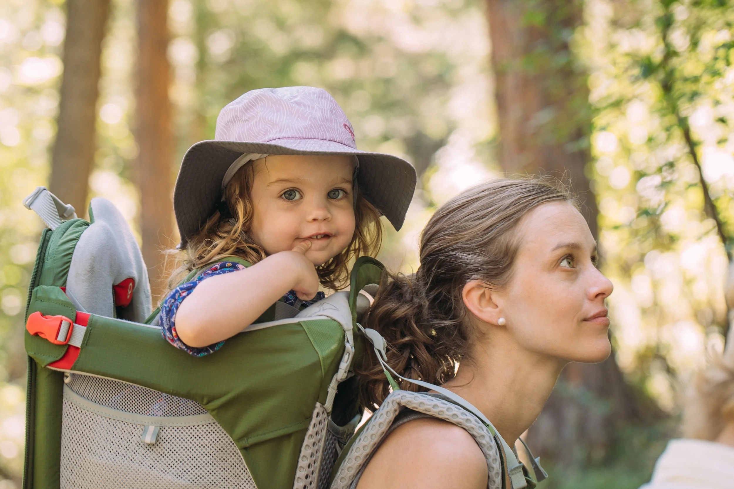 A woman carrying a young girl in a backpack carrier in a forest setting.