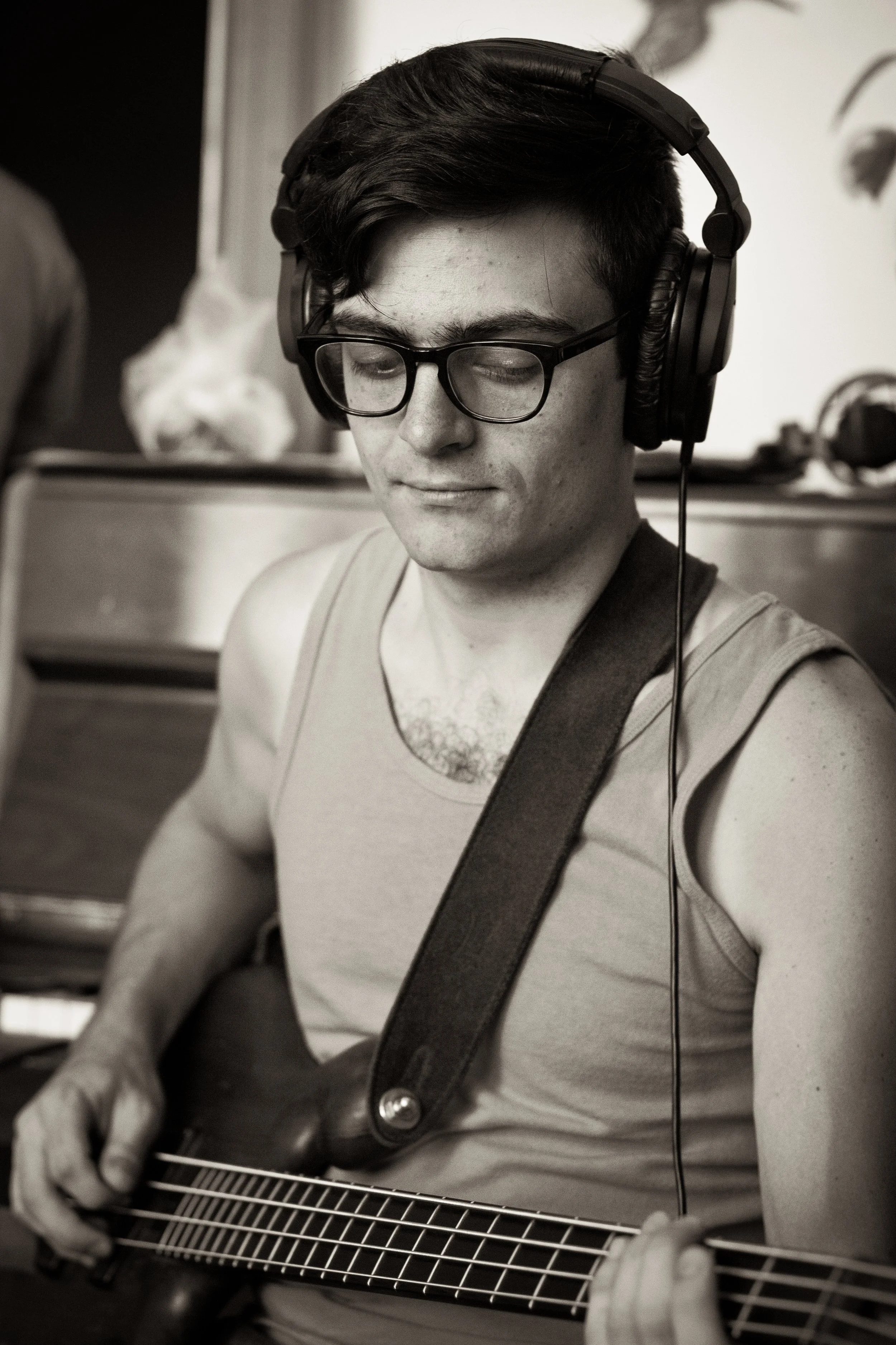 A young man with dark hair, glasses, and a sleeveless shirt playing an electric guitar, wearing large headphones.
