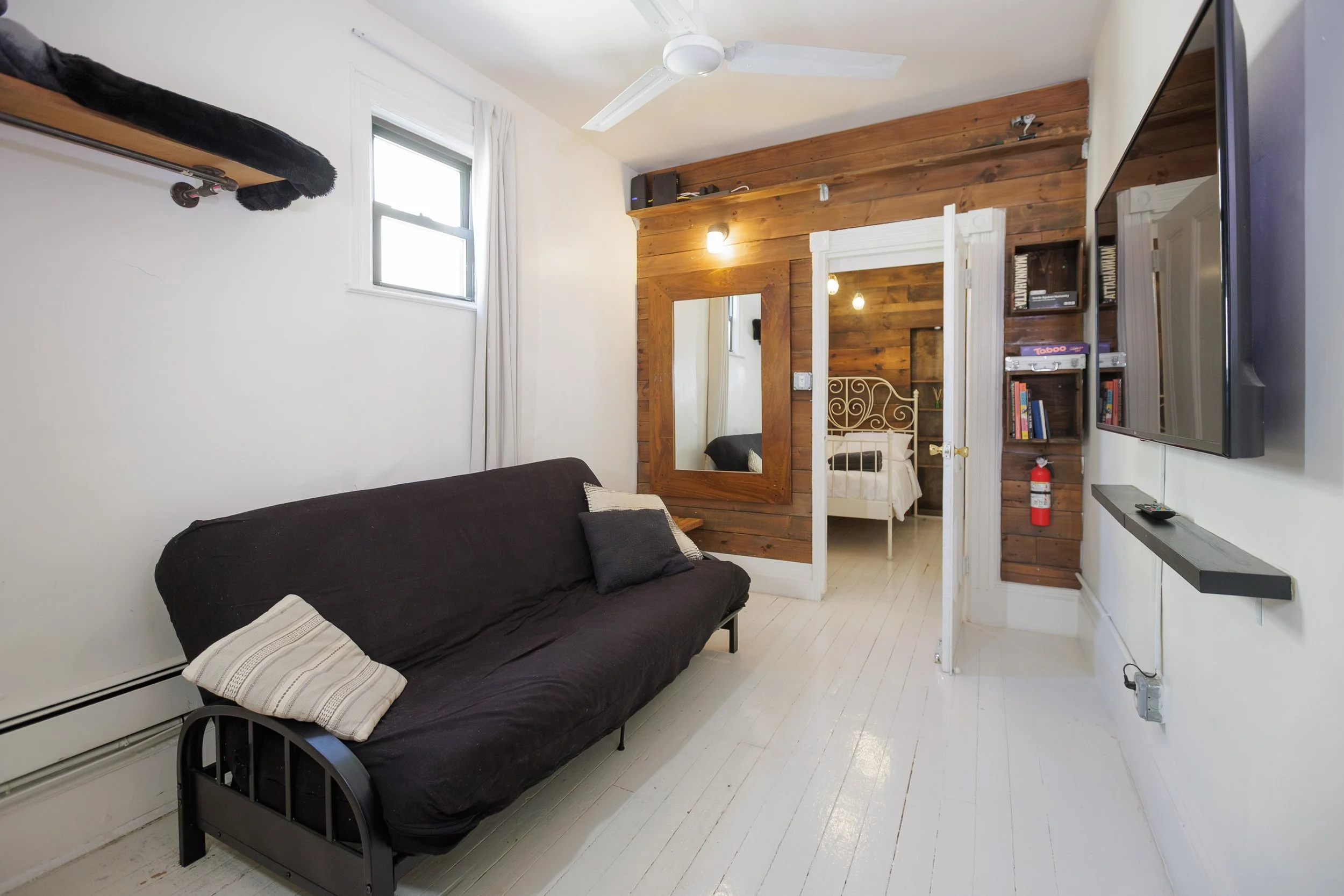 Small bedroom with a black futon on white wooden floor, white wall, window with white curtains, wooden accent wall with mirror, bed visible in the mirror, wall-mounted shelf and TV, fire extinguisher, ceiling fan.