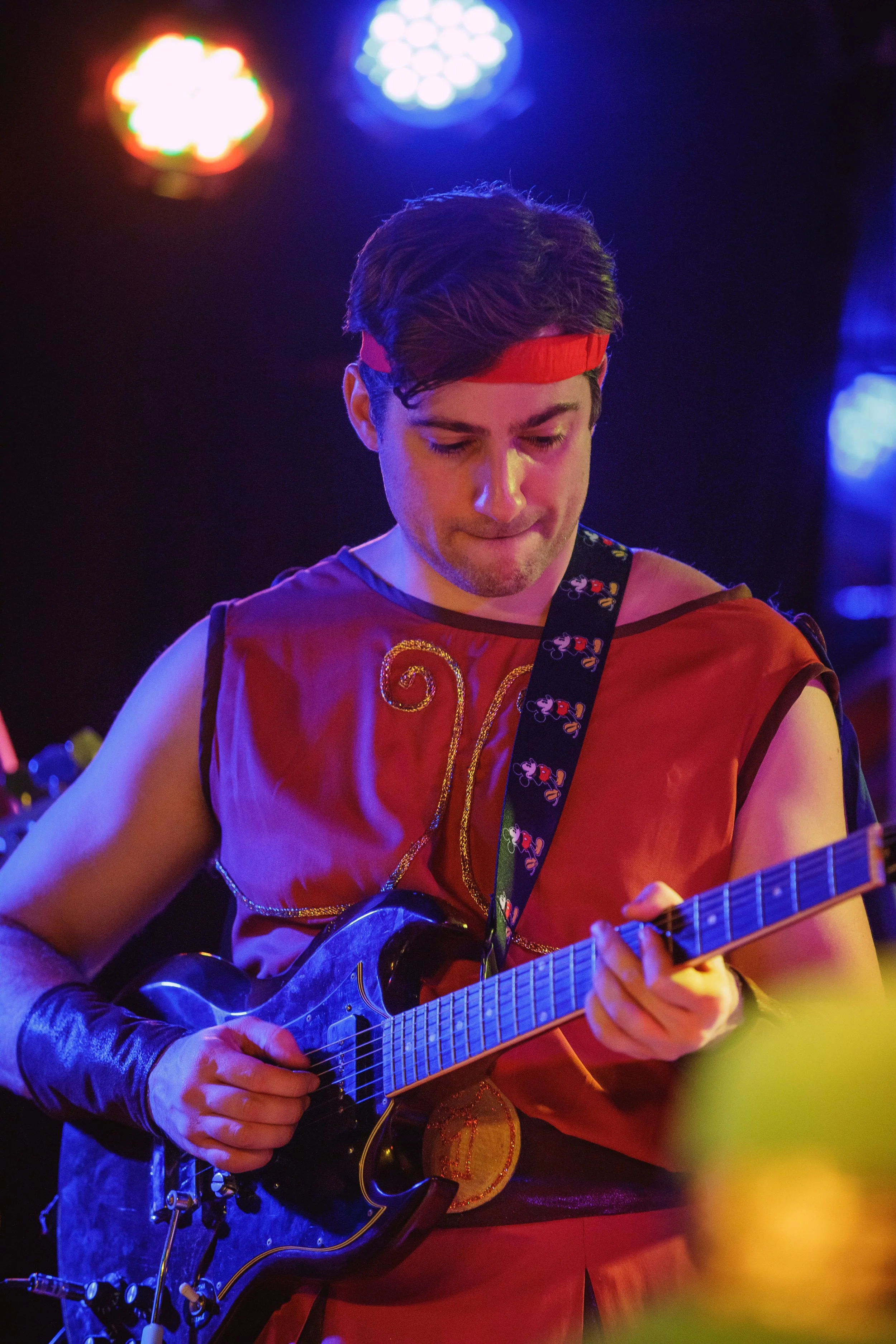 A young man in a red costume with gold embroidery, wearing a red headband, playing an electric guitar on stage under colorful lights.