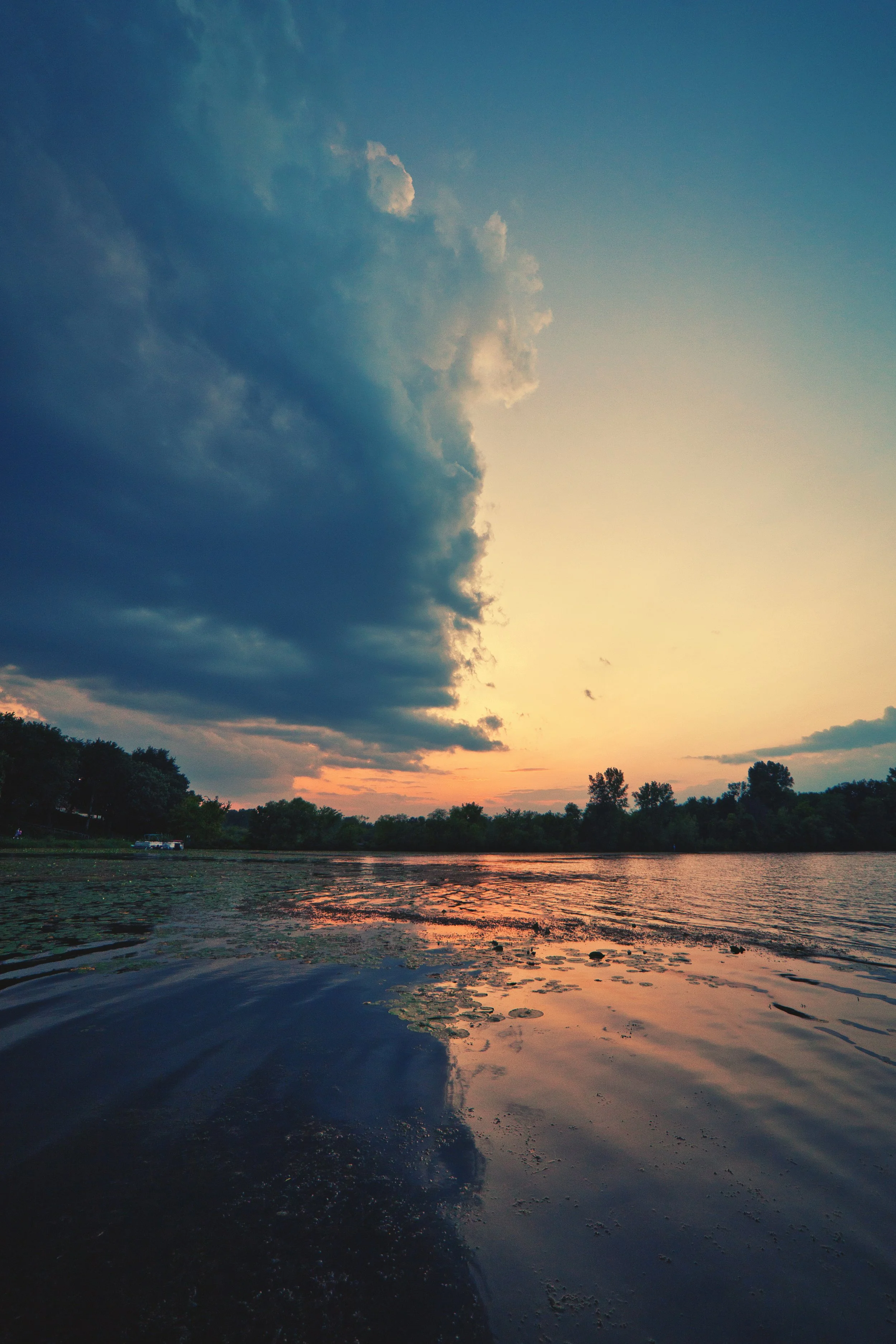 A tranquil river scene at sunset with the sky partially covered by dark clouds on the left and clear sky on the right, reflecting on the water with trees along the riverbank.