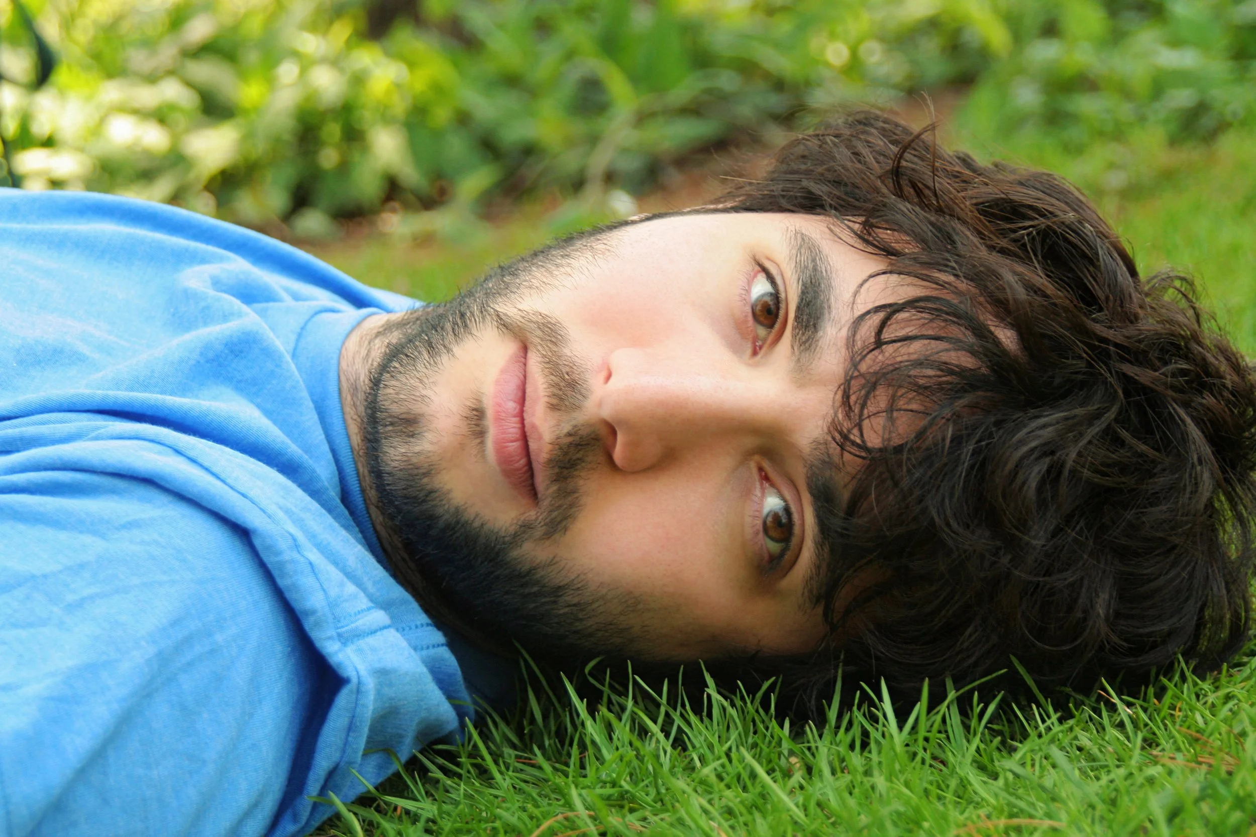 A young man with brown eyes, dark curly hair, and facial hair lying on green grass, wearing a light blue shirt, outdoors with blurred green foliage in the background.