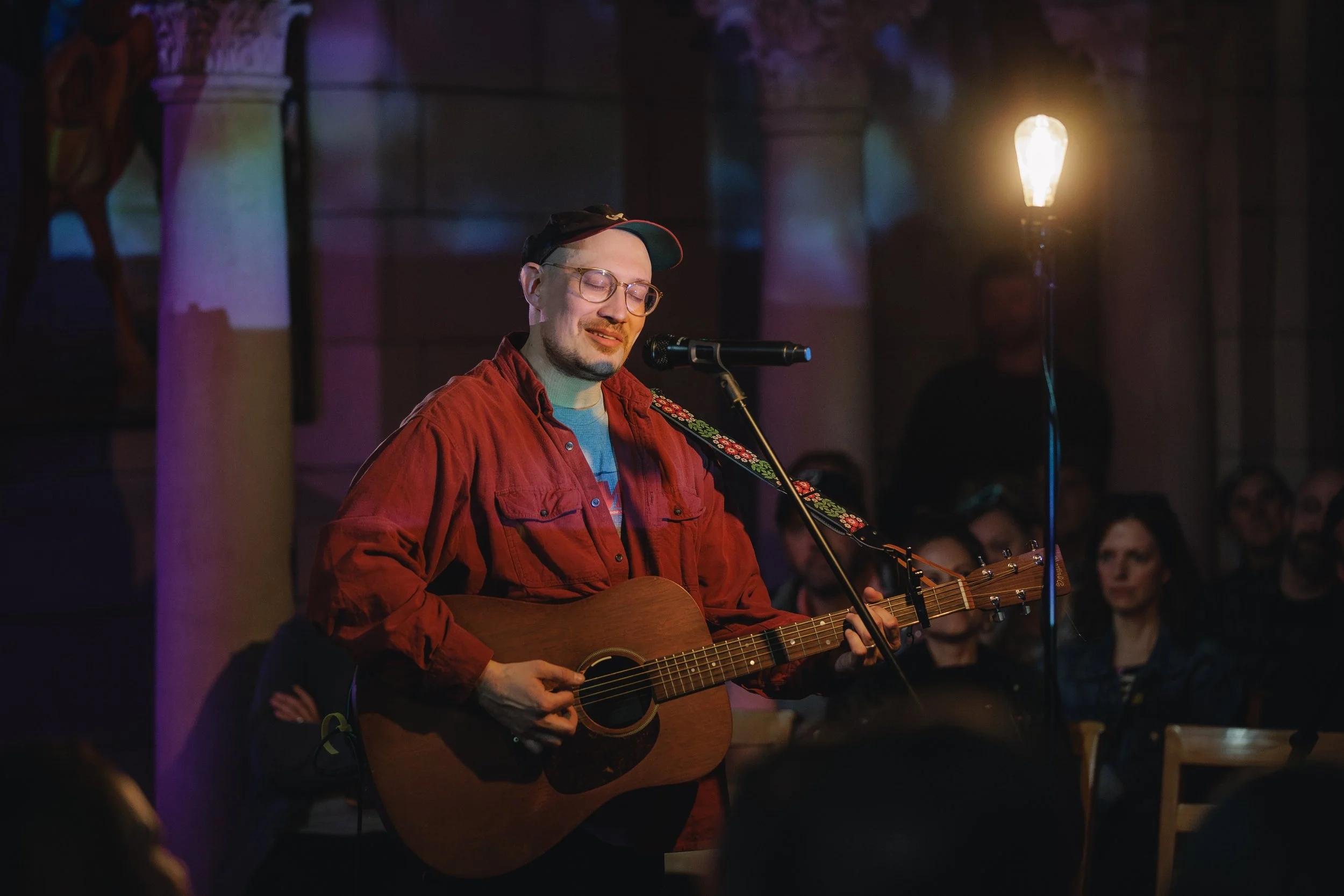 A man with glasses, wearing a red jacket and a baseball cap, is playing an acoustic guitar and singing into a microphone at a dimly lit indoor event. Several people are visible in the audience in the background.