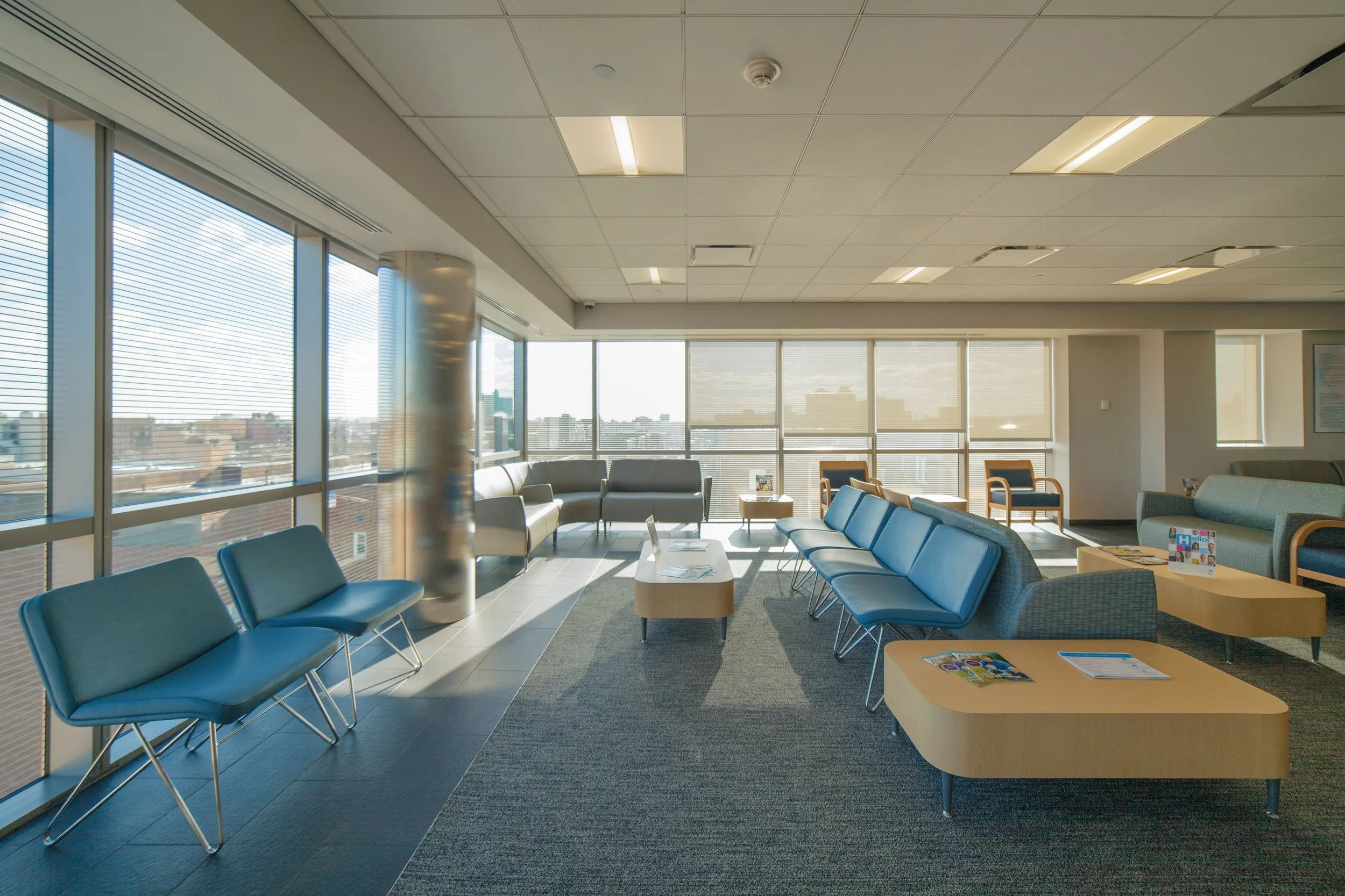 A modern office waiting lounge with large windows and city view, blue and gray seating arrangements, coffee tables, and reading materials on tables.
