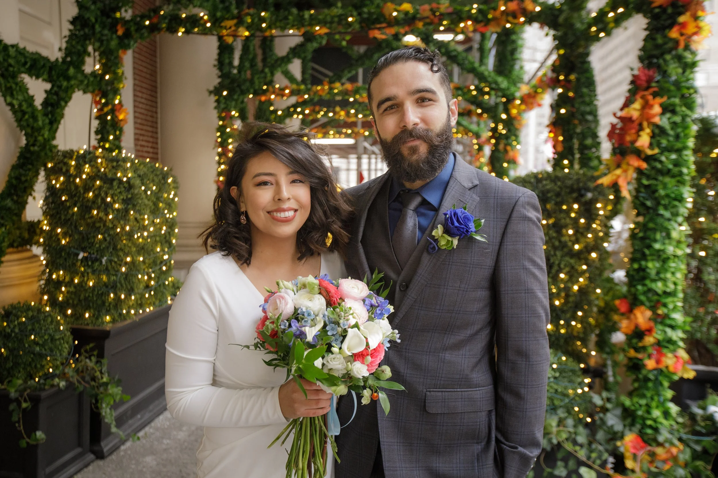 A newlywed couple standing close together in front of decorated greenery with lights, the bride holding a bouquet and smiling, the groom dressed in a suit with a boutonniere, both smiling warmly.