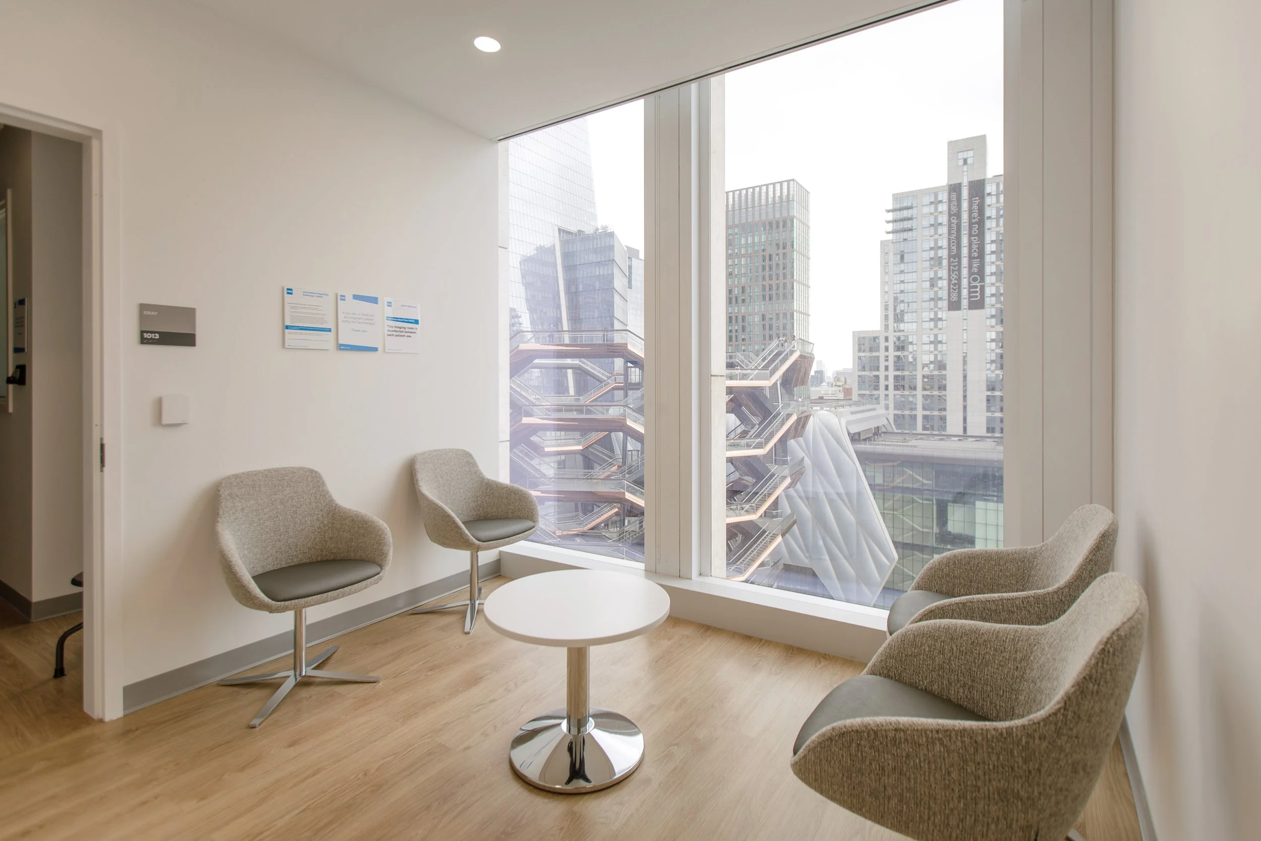 A modern office waiting area with four beige armchairs around a small round white table, large windows showing city buildings, and a wall with informational posters.