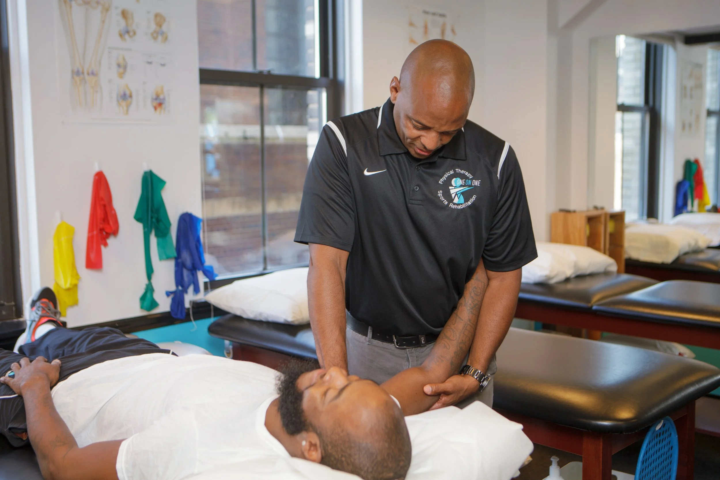 A healthcare professional provides physical therapy to a patient lying on a treatment table, with medical posters on the wall and exercise bands hanging in the background.