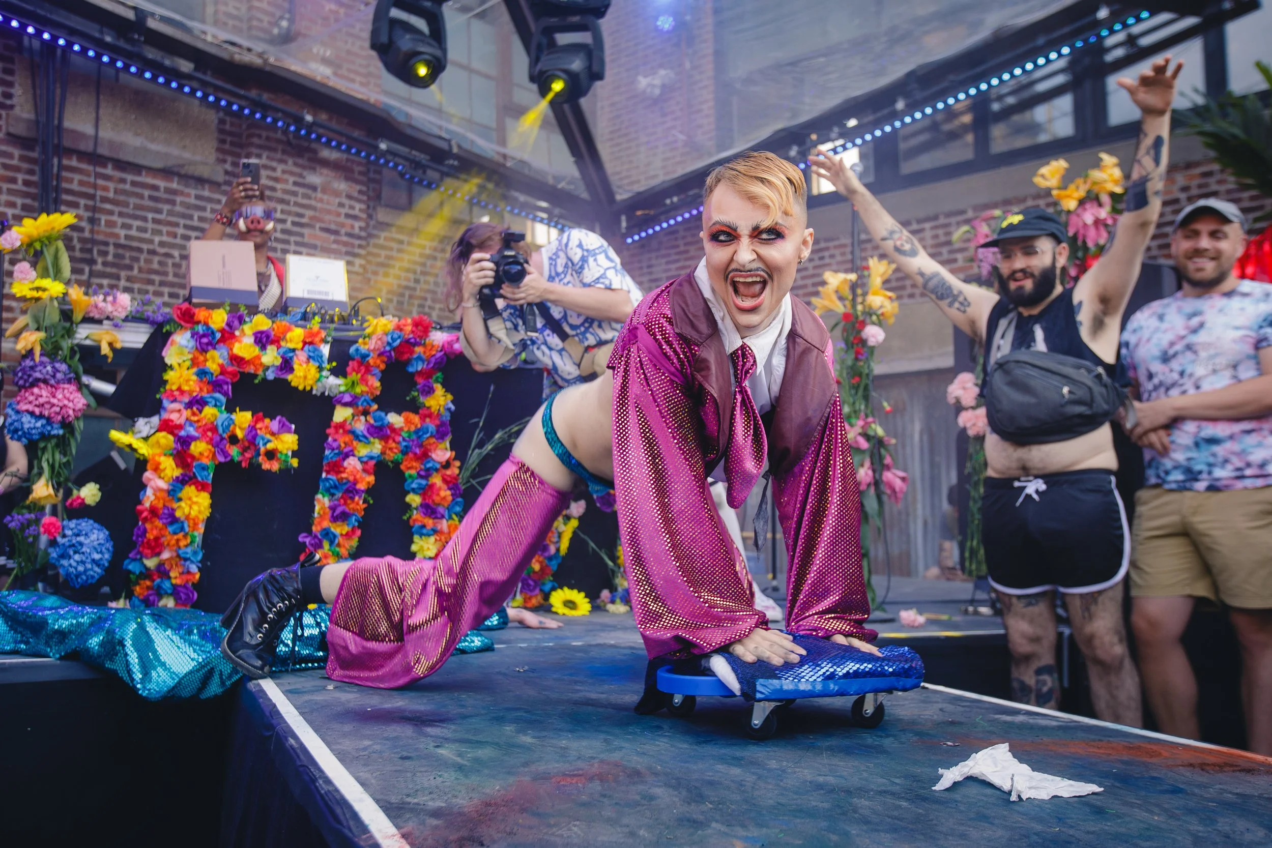 A performer in a pink and purple outfit crawls on the stage during a lively event with a floral backdrop, other people, a photographer, and colorful stage decorations.