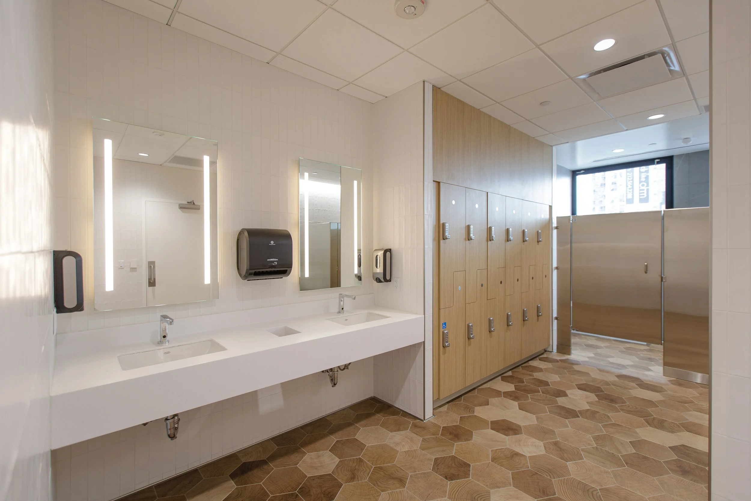 Public restroom with two sinks, large mirror, soap dispenser, hand dryer, wooden lockers, and hexagonal tile flooring.