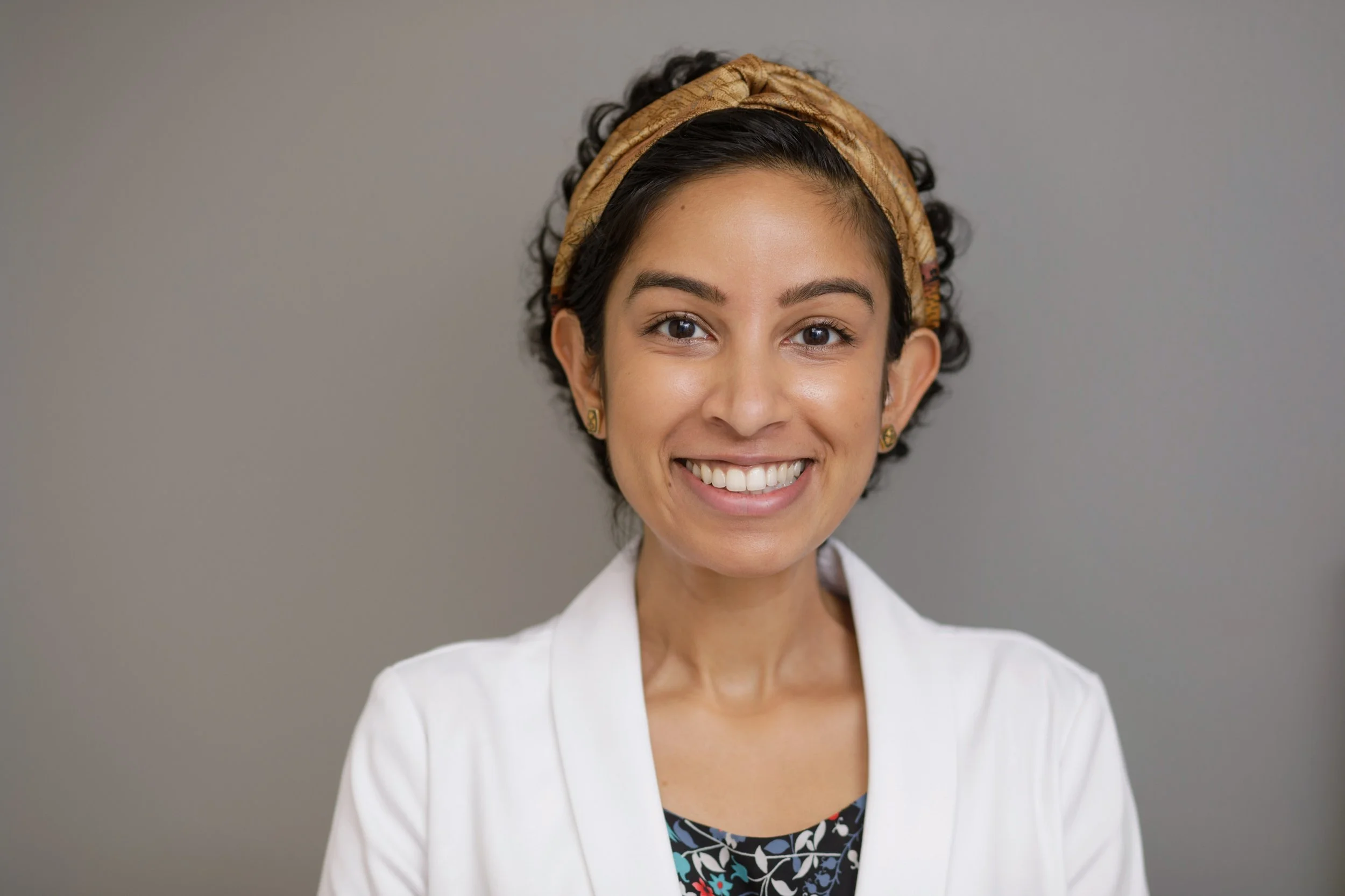 A close-up portrait of a woman with dark, curly hair styled with a tan headband, wearing earrings, and smiling with visible teeth against a neutral gray background.
