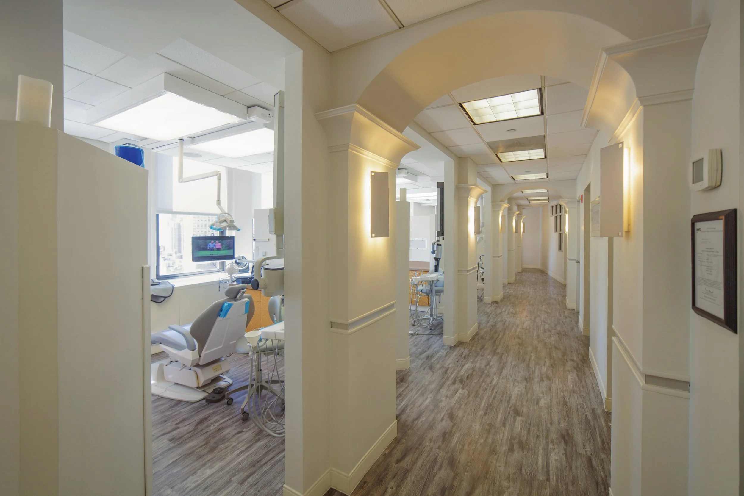 Hallway in a dental clinic with white walls, wood flooring, and ceiling lights. On the left, a treatment room with dental chairs, medical equipment, and a window with city view.