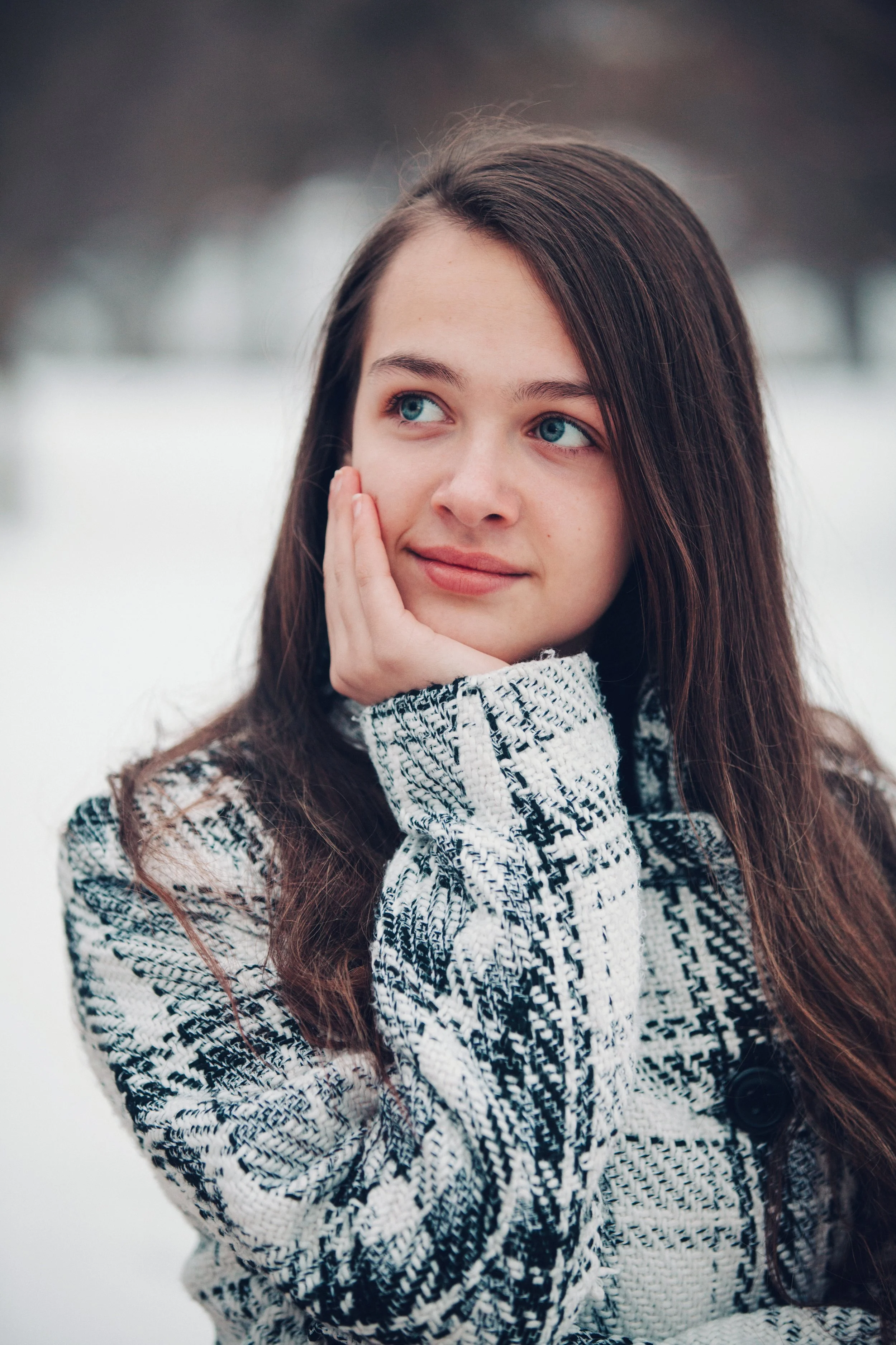 A young woman with blue eyes and long brown hair outdoors in a snowy setting, wearing a black and white patterned jacket and resting her face on her hand.
