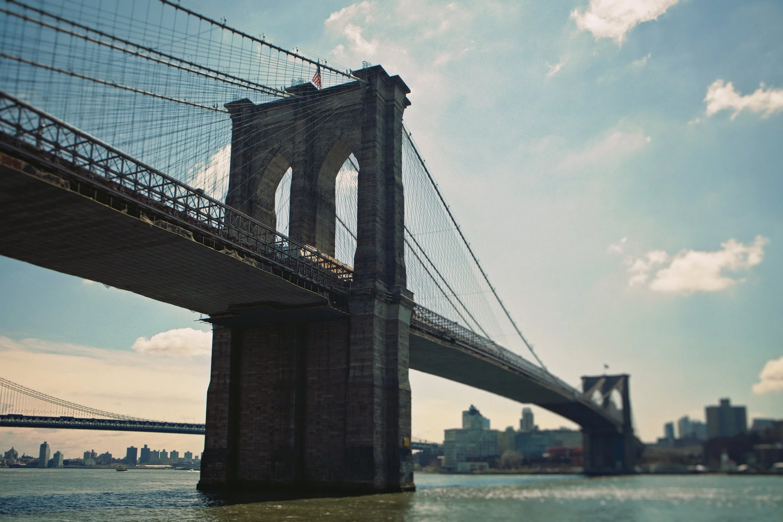 The Brooklyn Bridge with its stone towers and suspension cables, spanning over the East River in New York City, with city skyline in the background and a partly cloudy sky.