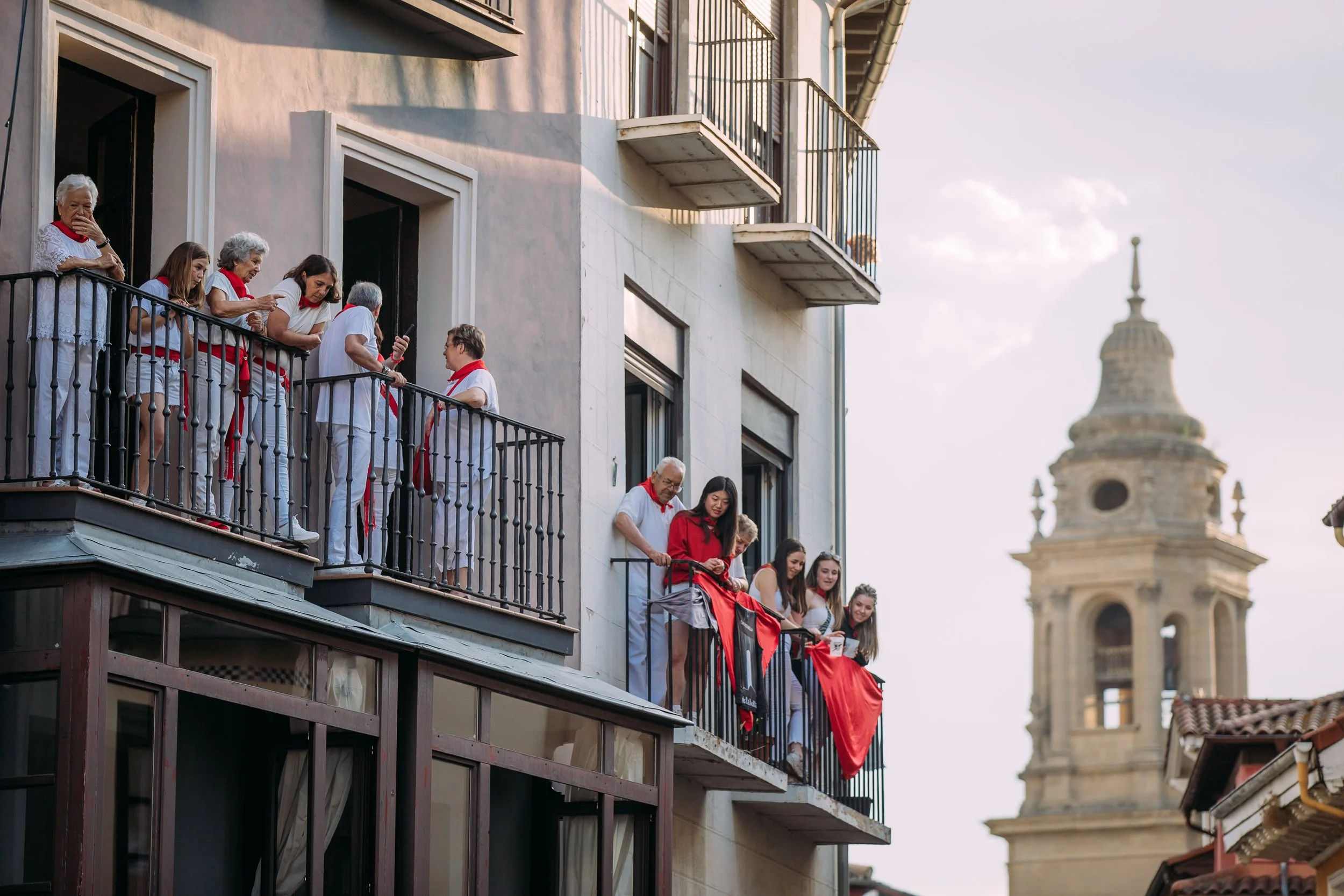 Several people are gathered on balconies of a building overlooking a street. Some are dressed in white with red scarves, indicating participation in a celebration or festival. A church tower is visible in the background, suggesting a European city se