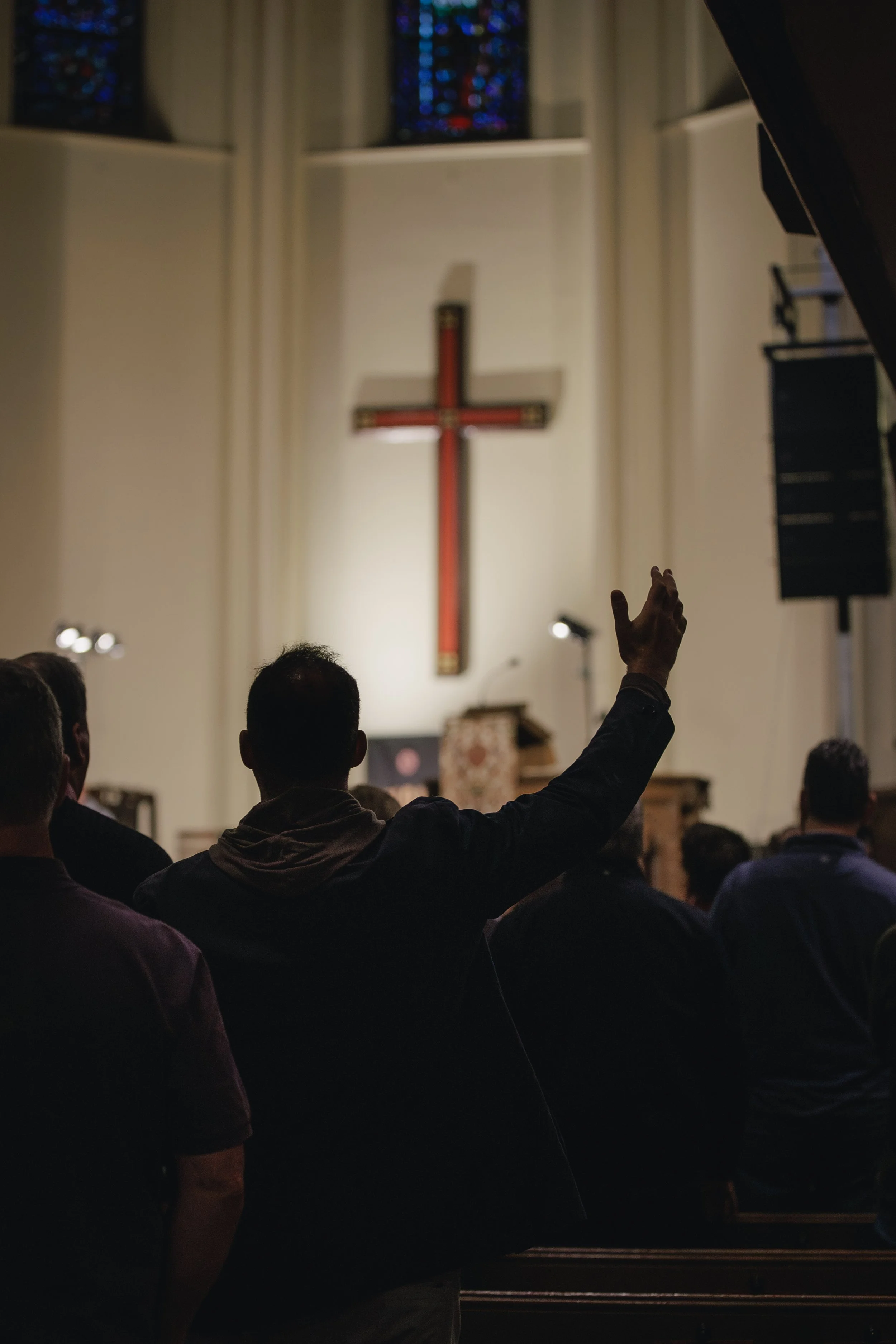 People sitting inside a church, with a person raising their hand, and a large cross on the wall in the background.