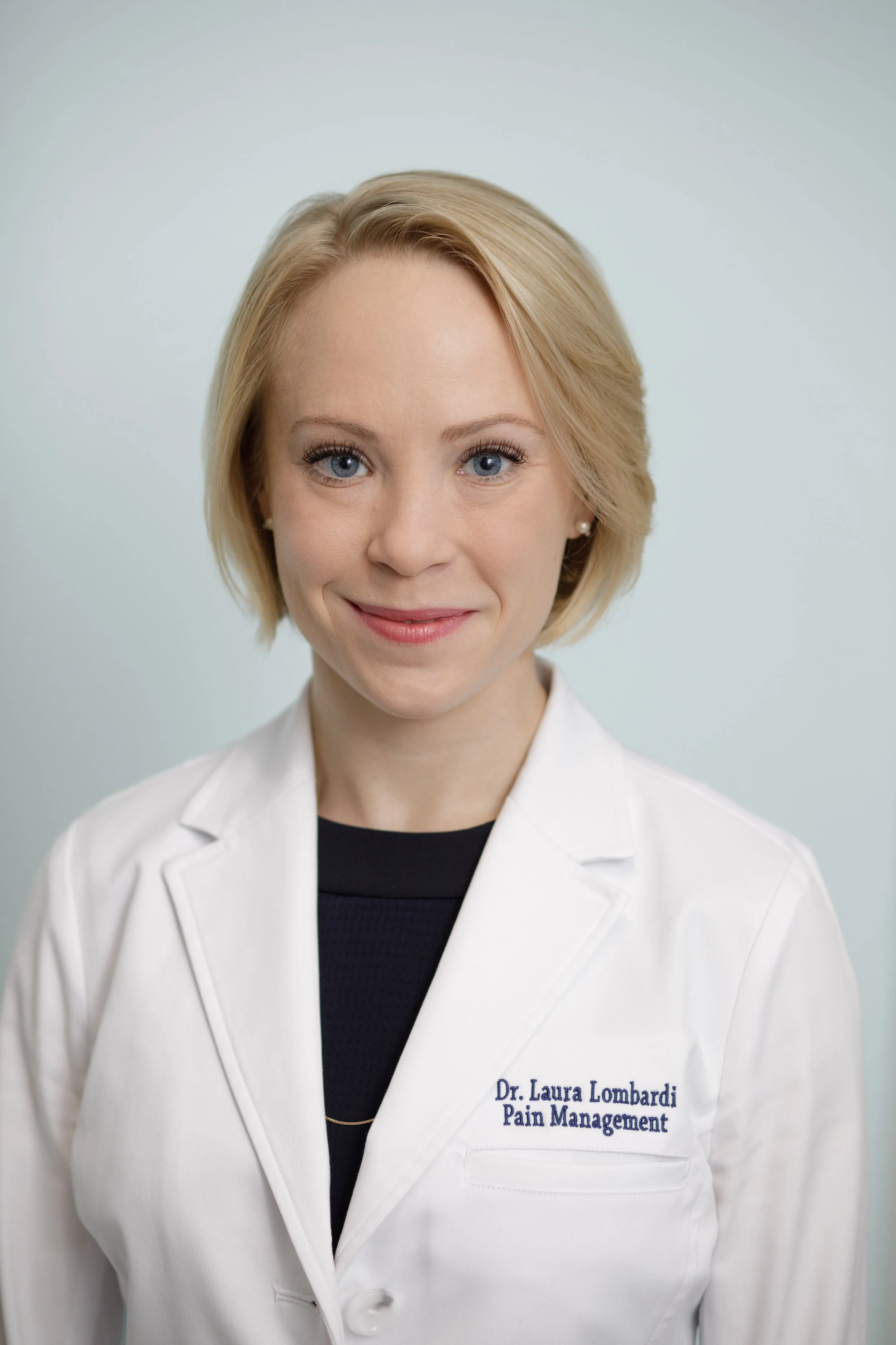 A woman with blonde hair wearing a white medical coat with embroidered text 'Dr. Laura Lombardi Pain Management', a black top underneath, and pearl earrings, standing against a plain light background.