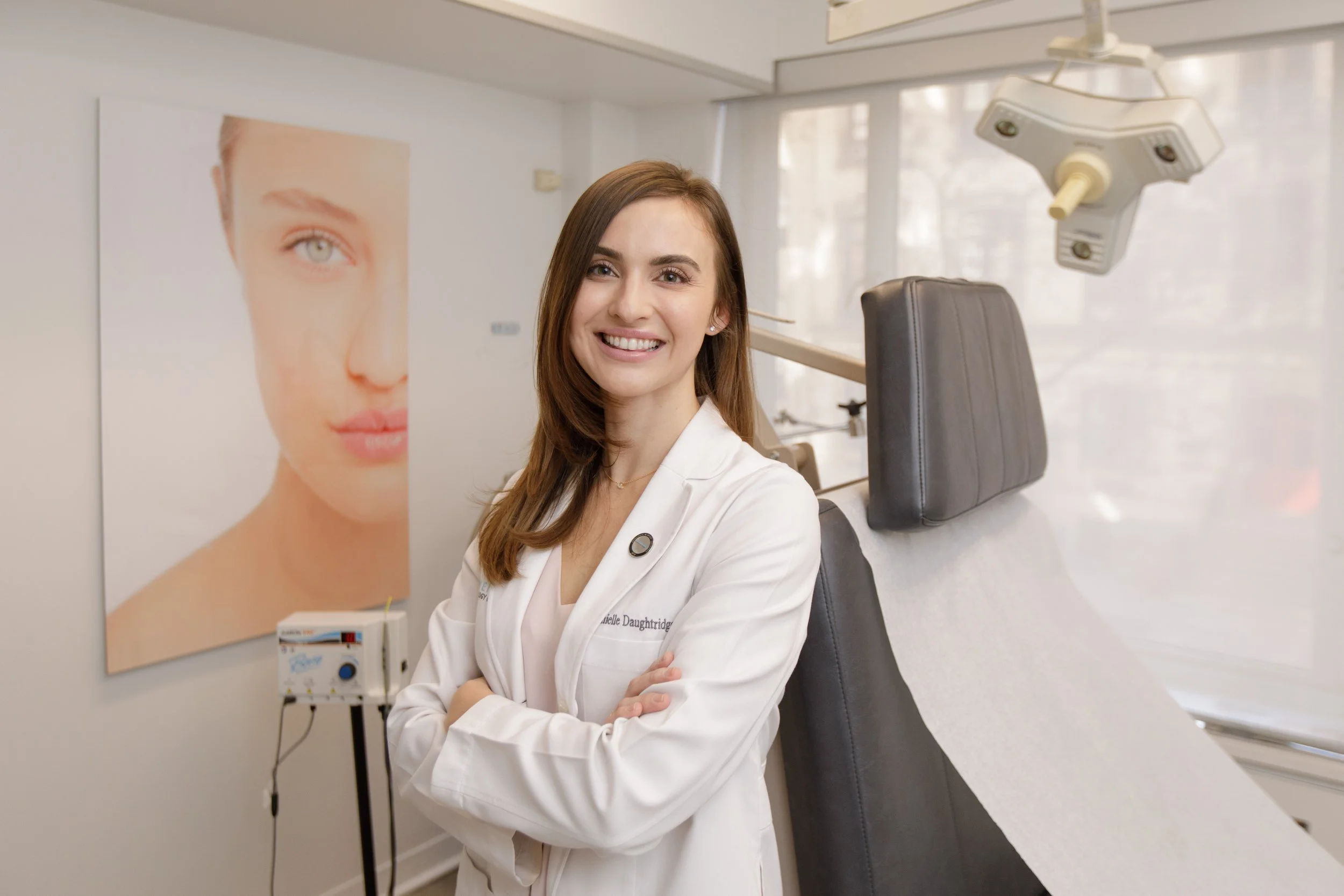 A female dentist with long brown hair, wearing a white lab coat, smiling and standing with arms crossed in a dental office.