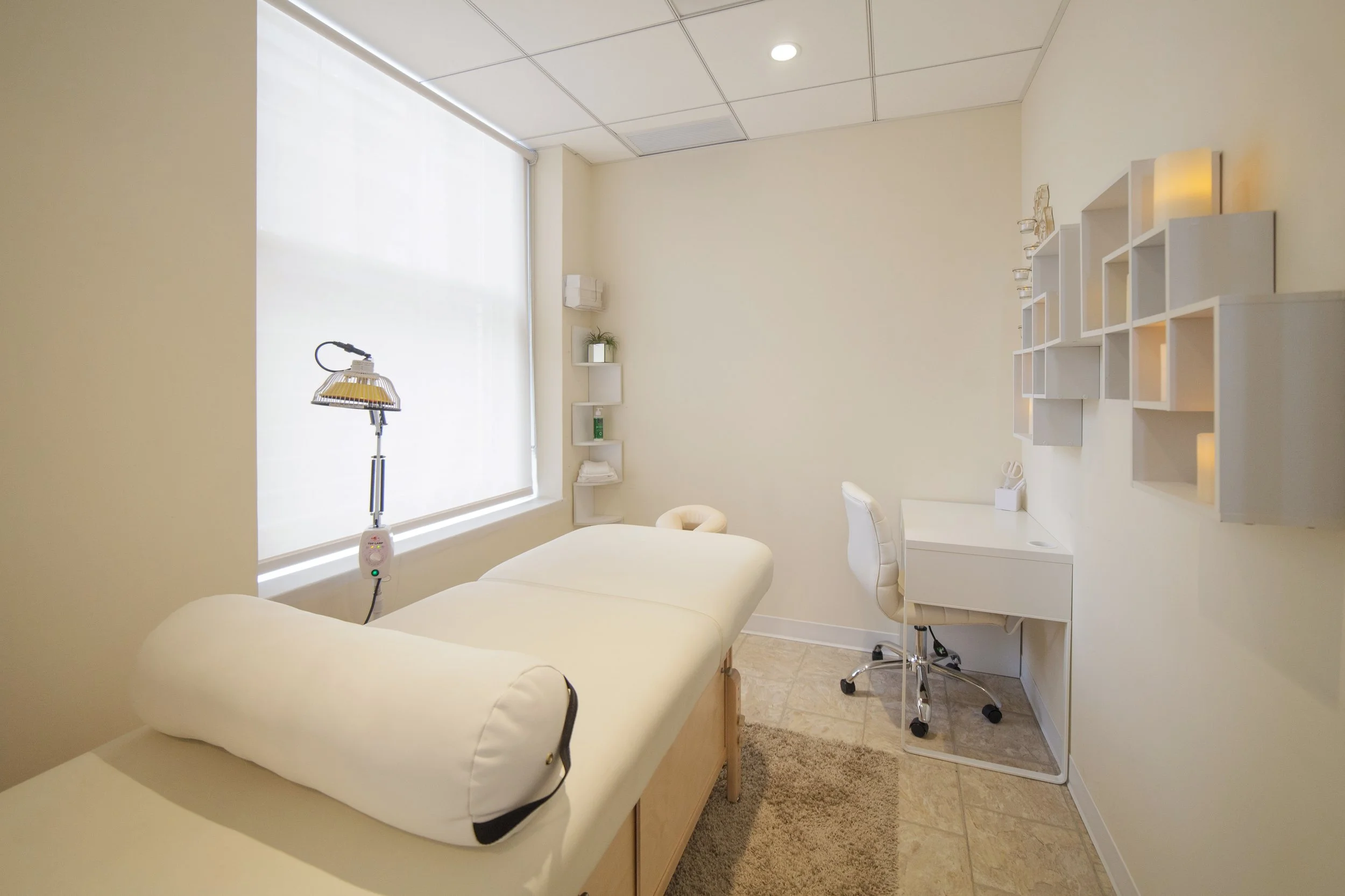 A clean, bright medical examination room with a massage or treatment table, white walls, a window with sheer blinds, and minimal decor including shelves with small items and a potted plant. There is a lamp and a rolling stool in the room.