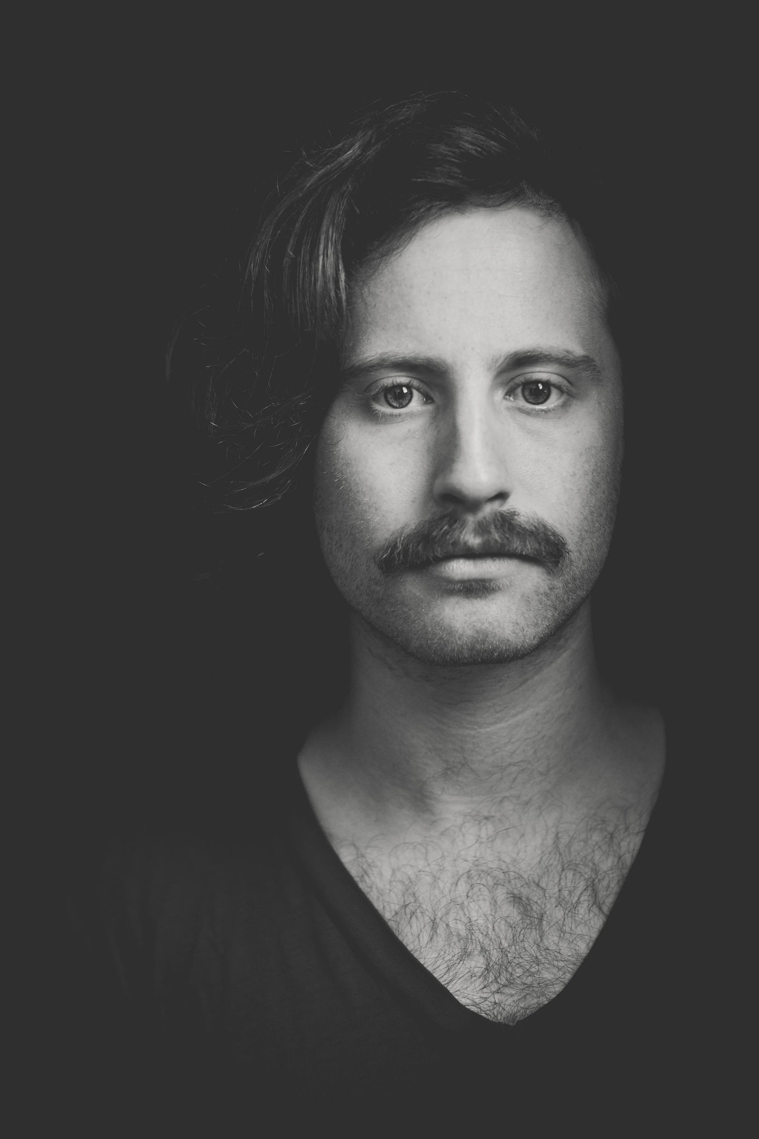 Black and white close-up portrait of a man with wavy hair, a mustache, and a beard, wearing a dark shirt, against a dark background.