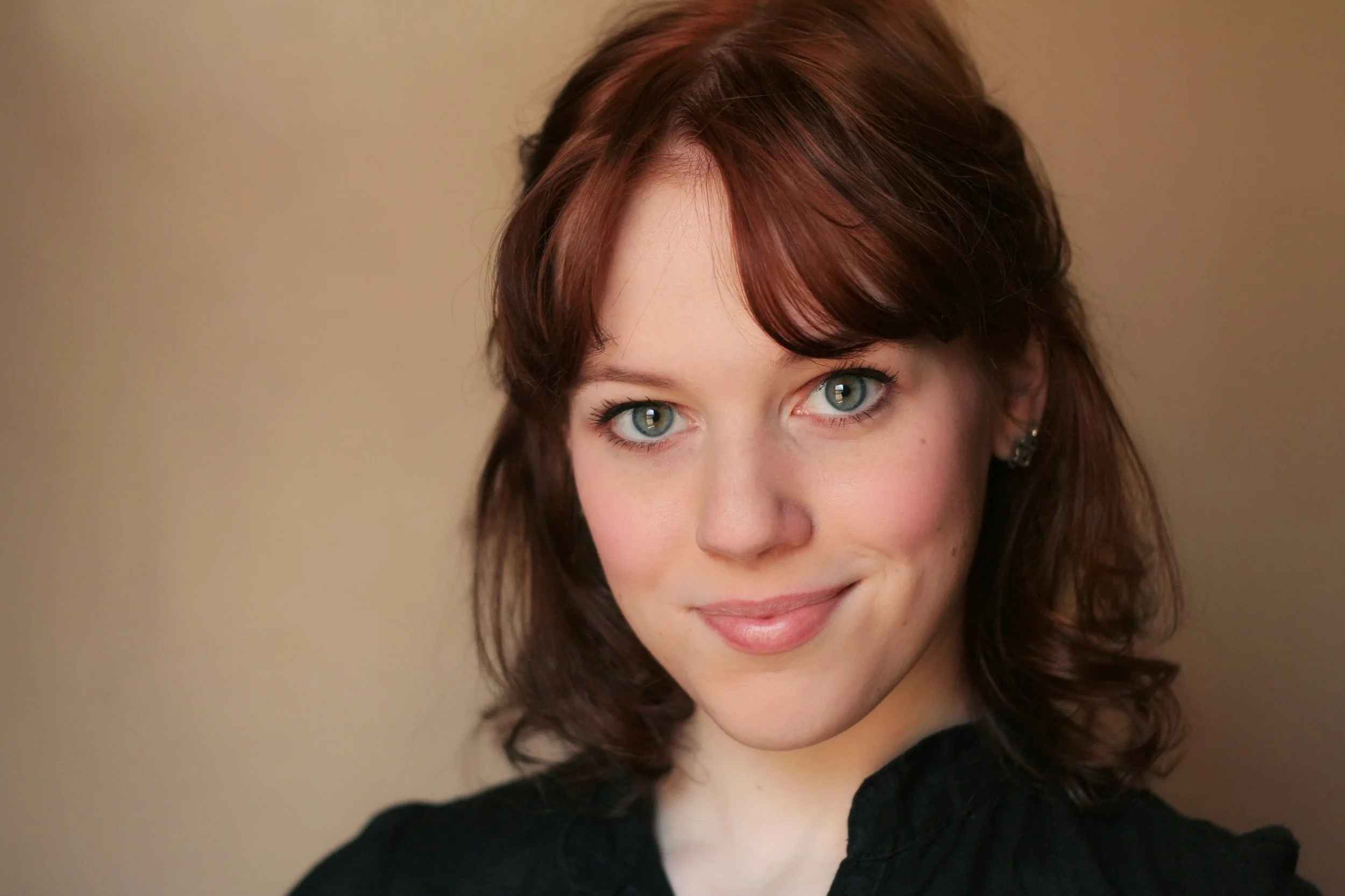 Close-up portrait of a woman with blue eyes and reddish-brown hair, wearing a black top and small earrings, smiling subtly. The background is plain and beige.