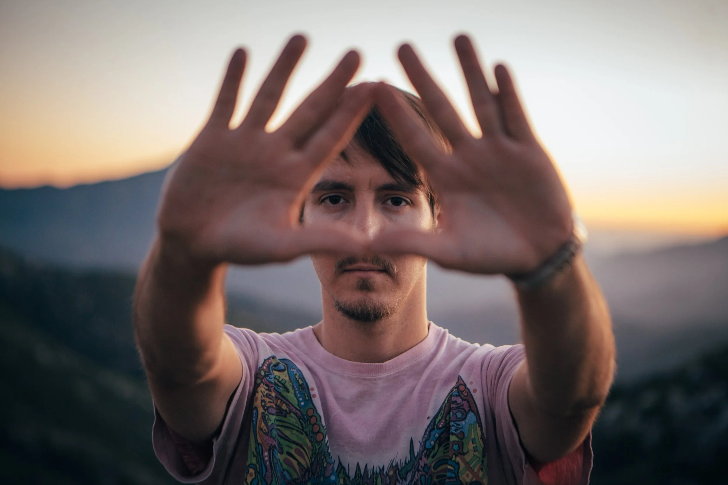 A young man with dark hair and facial hair is holding his hands in front of his face, forming a triangle shape, as he gazes directly into the camera with a sunset and landscape in the background.