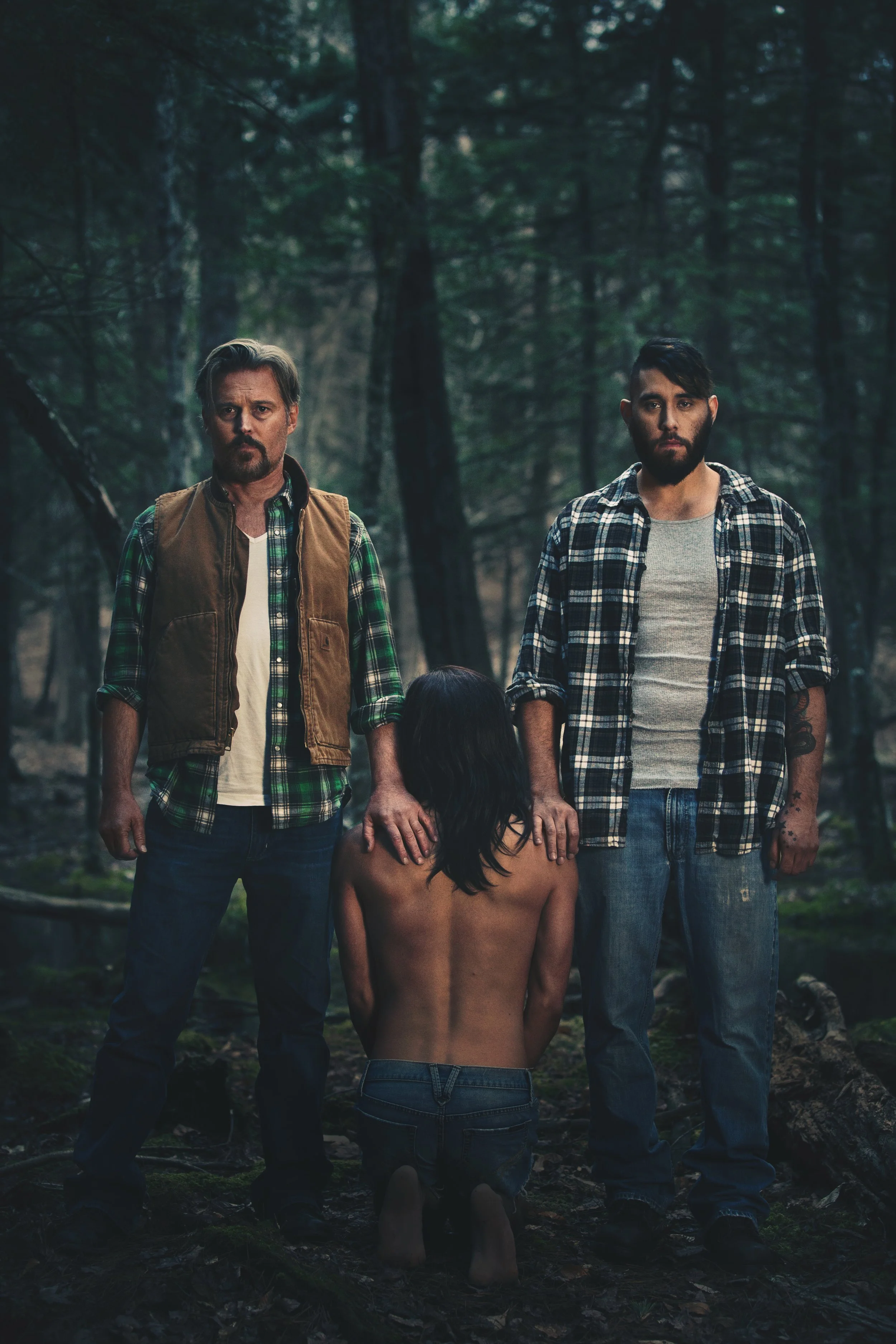 Two men stand in a dark forest holding hands with a naked woman kneeling in front of them, facing away from the camera.