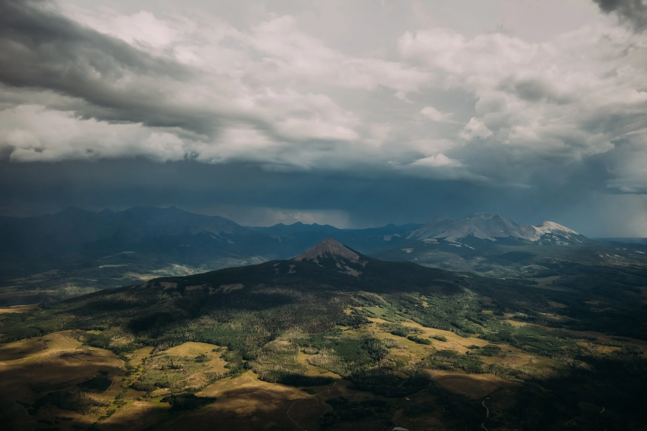 A landscape of mountains with dark storm clouds overhead. The foreground shows green and brown fields and a forest, leading to distant mountain ranges with patches of snow.
