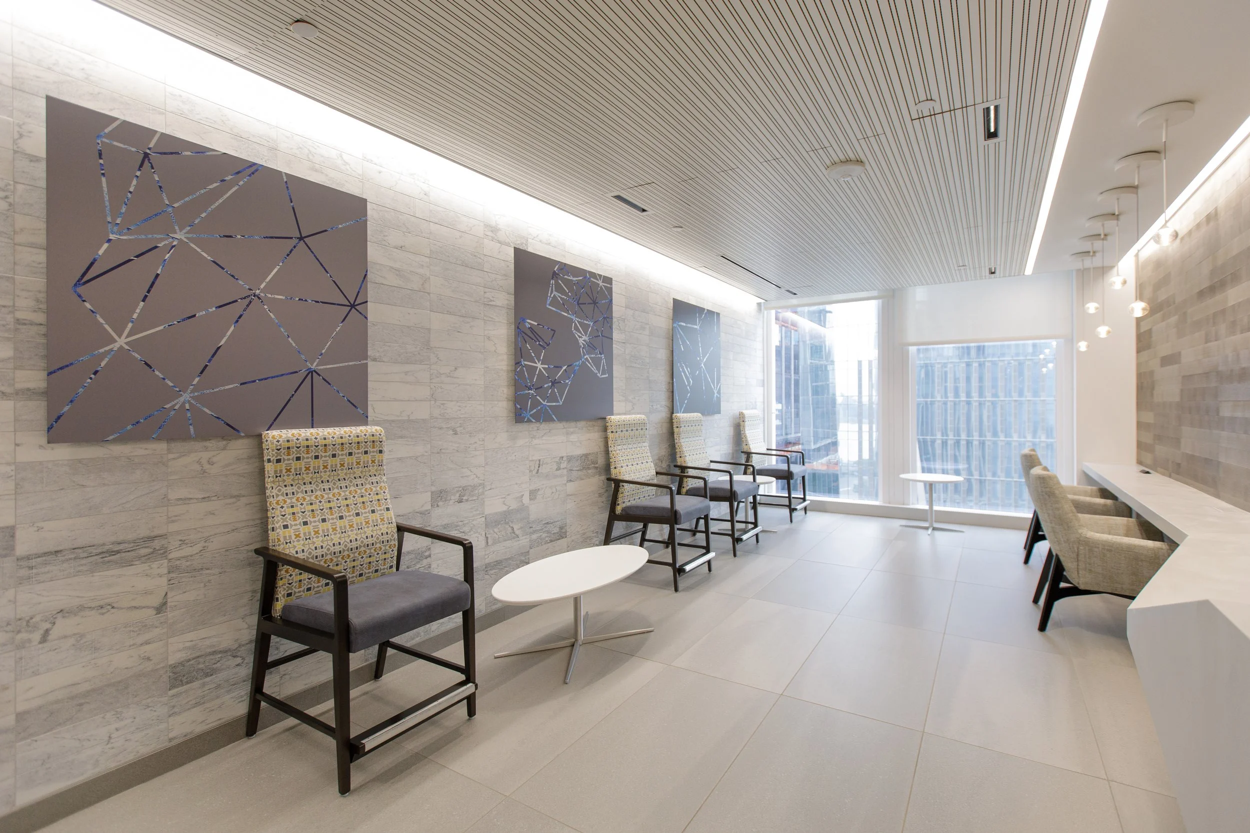 Interior of a modern lounge area with light-colored tiled floors and walls, wood-paneled ceiling, and large windows letting in natural light. There are patterned chairs, small round tables, and abstract artwork on the wall.