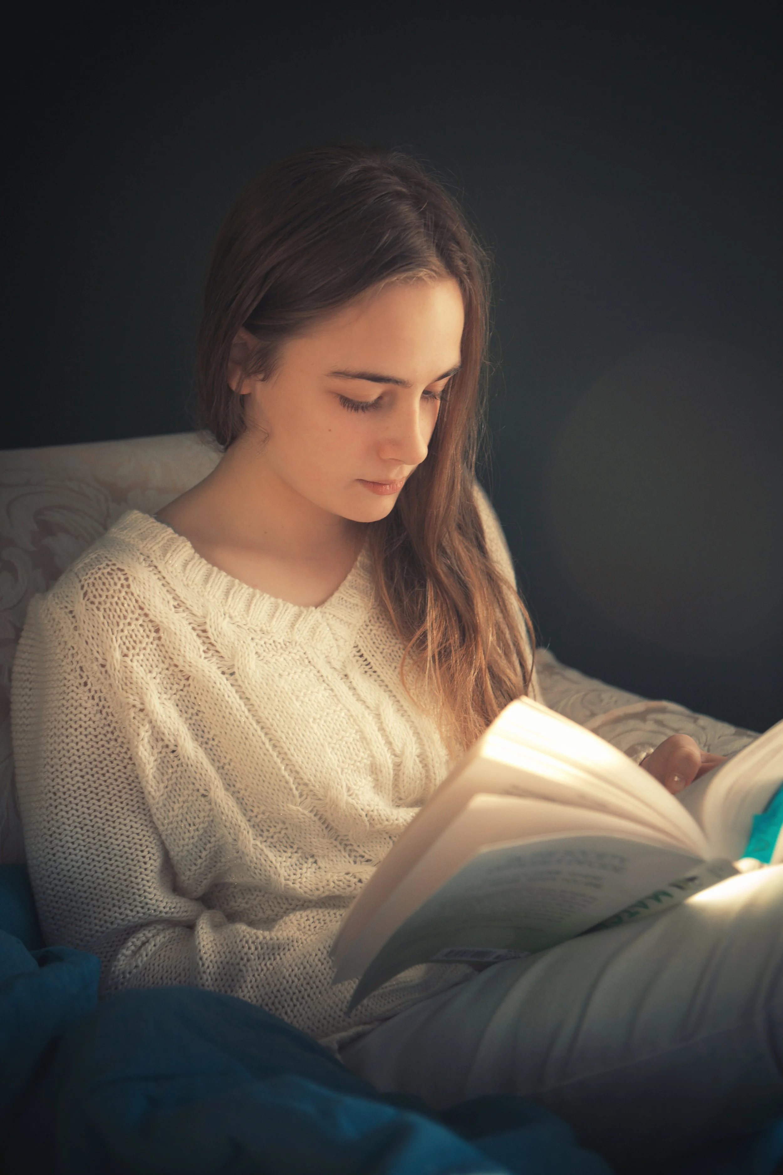 A young woman with long brown hair, wearing a cream-colored knitted sweater, is sitting in a dark room, reading a book.