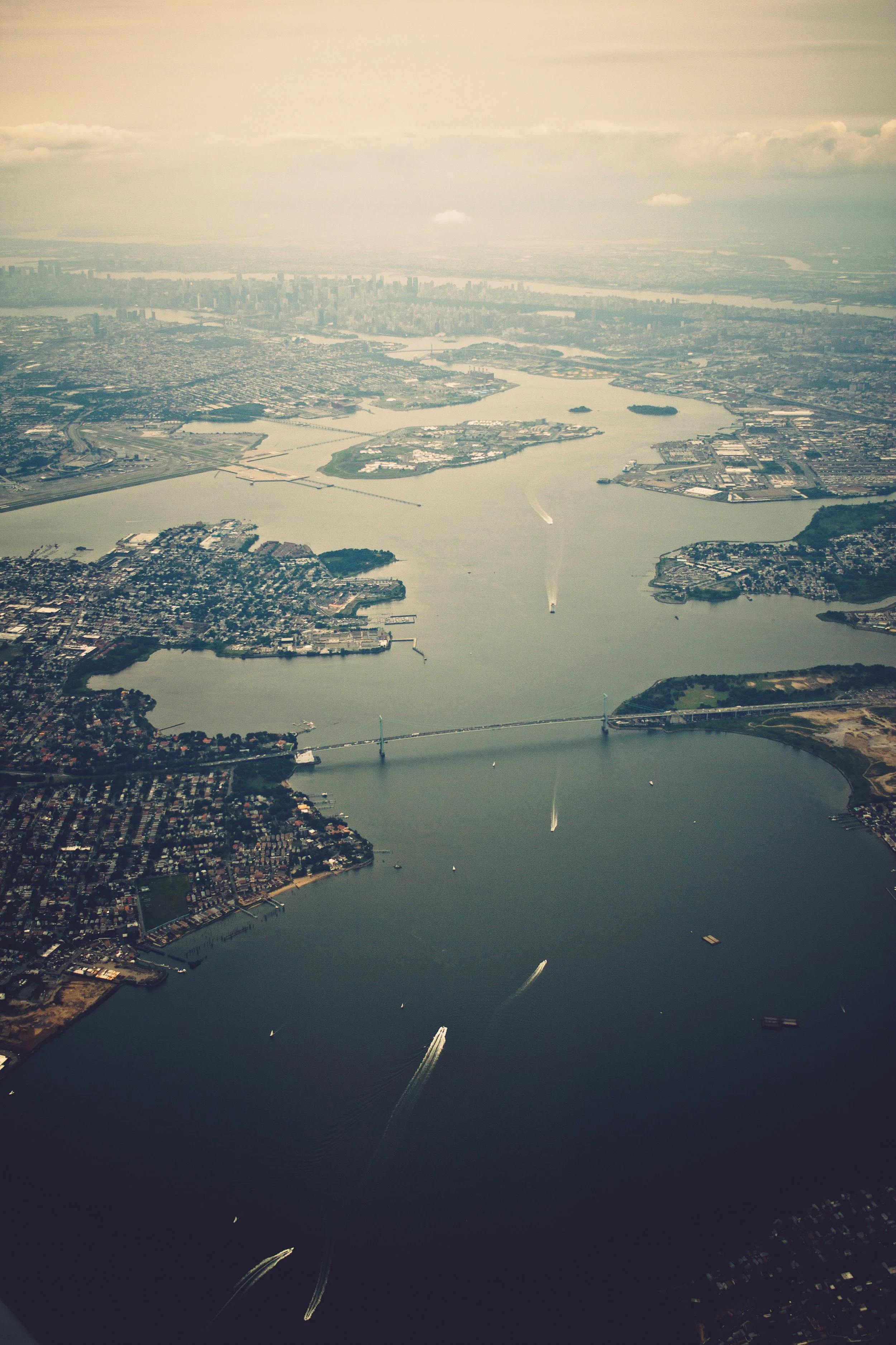 An aerial view of the New York City skyline and surrounding waterways on a cloudy day, with boats creating trails in the water.
