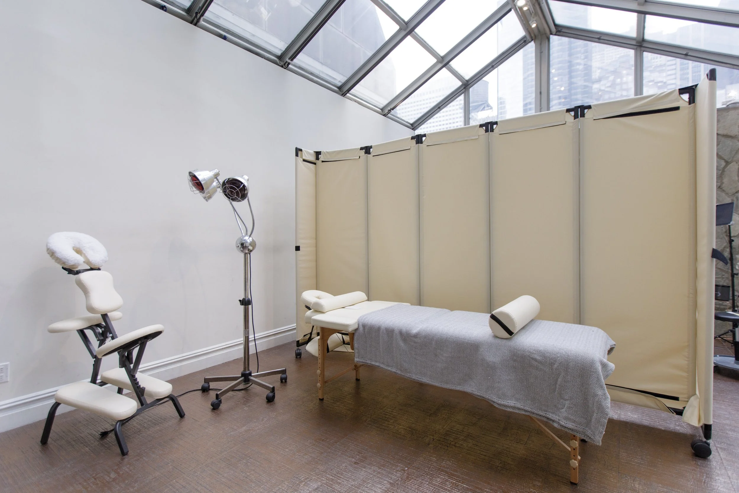 A medical or massage treatment room with a treatment bed covered in gray fabric, a beige privacy screen, a white stool, a small white stool, a lamp with two adjustable heads, and a large window with a view of tall buildings. The room has brown floori