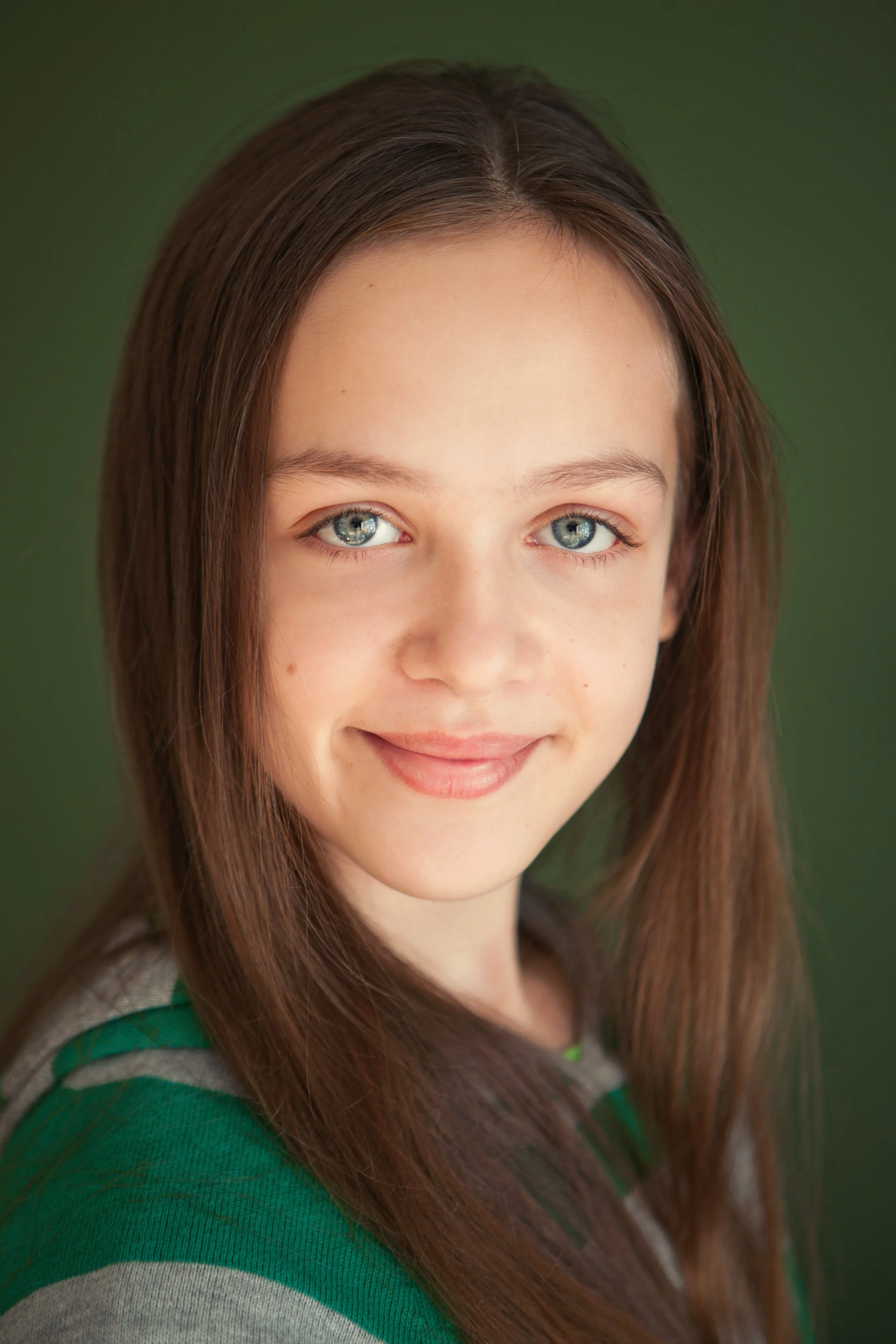 Close-up of a smiling young woman with long brown hair, blue eyes, wearing a green and gray striped shirt, against a green background.