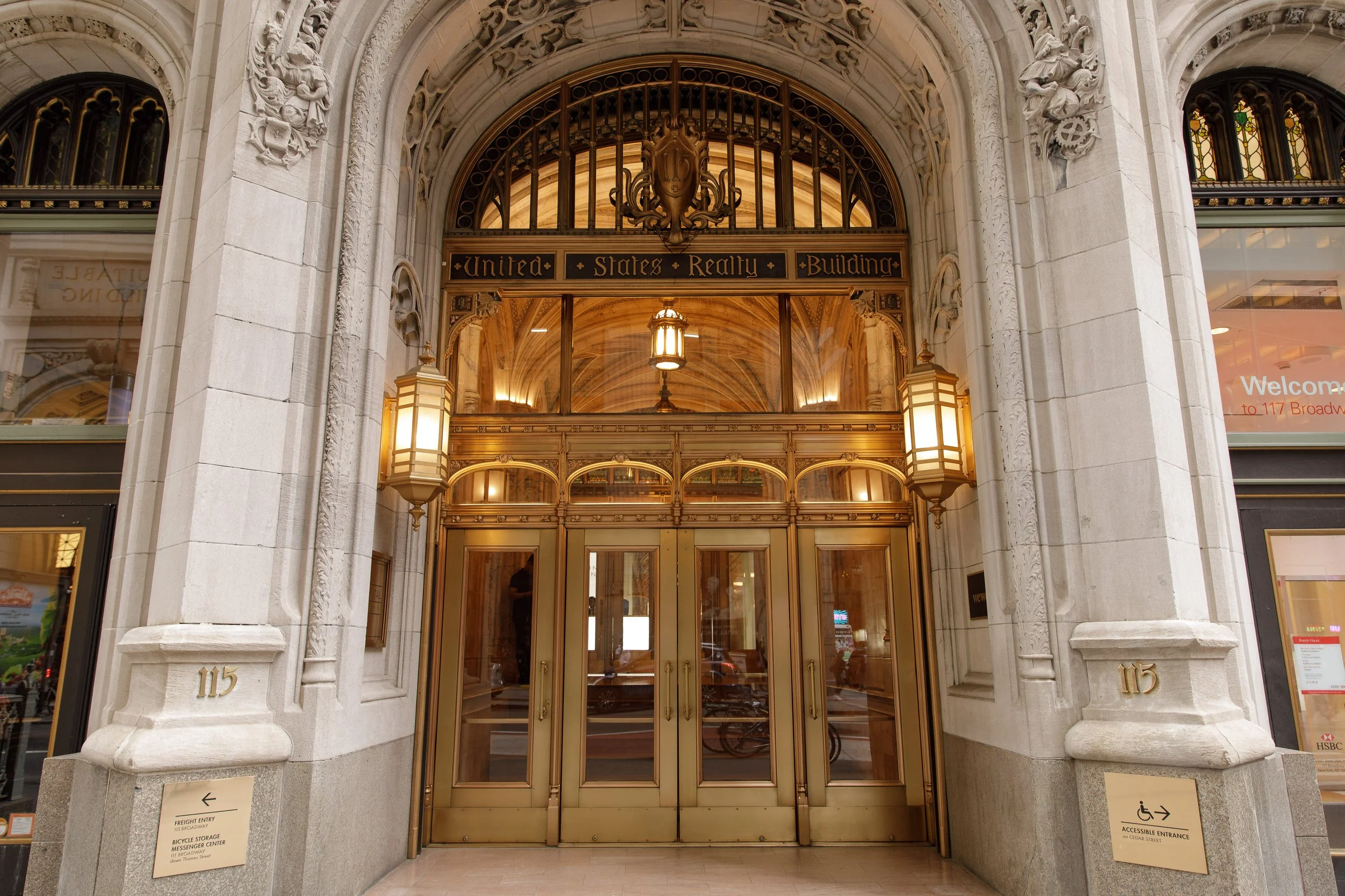 Entrance to the United States Realty Building with ornate architectural details, golden doors, and lighting fixtures. Signage indicating it is at 115 Broadway, with accessible entrance signage nearby.