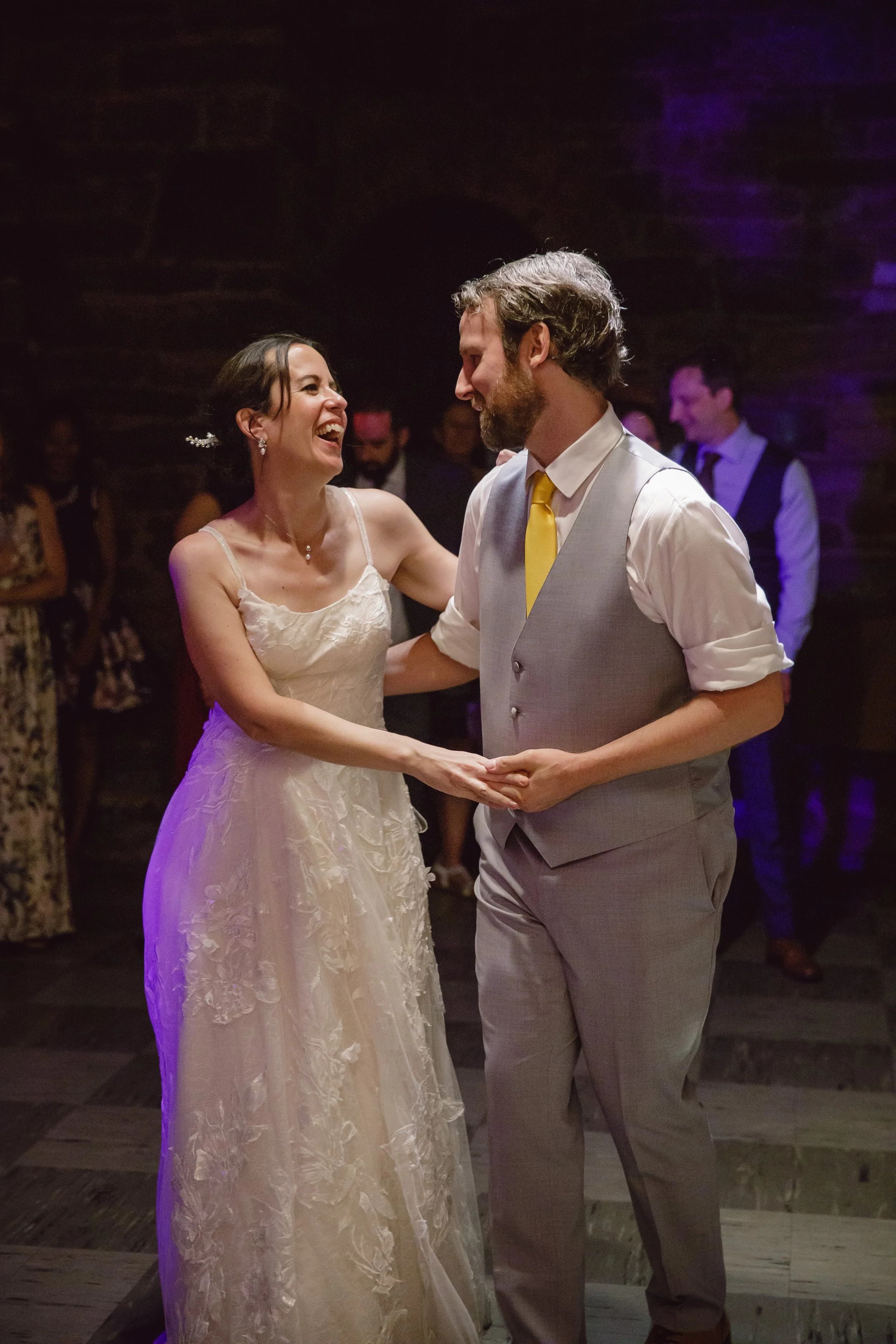 A bride and groom share a happy dance at their wedding reception. The bride is wearing a white lace wedding gown, and the groom is dressed in a light gray vest with a yellow tie. They are smiling and looking at each other, surrounded by guests in a d