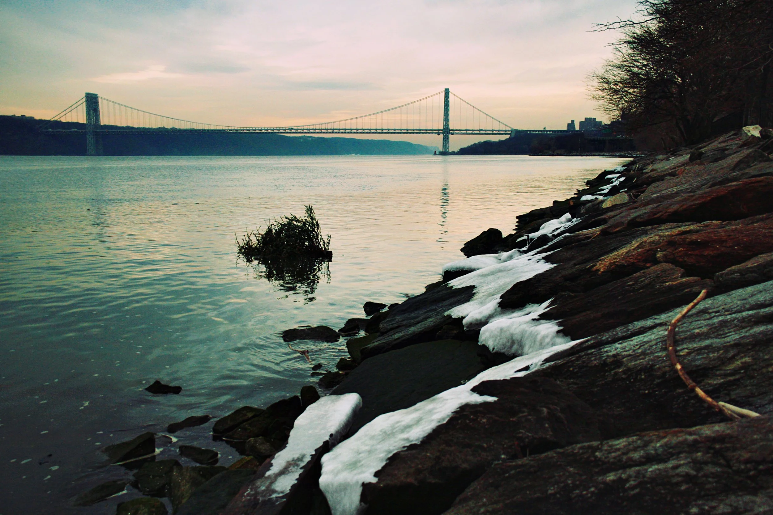 View of a suspension bridge over a river with rocky shoreline and snow patches, trees on the right, and cloudy sky.