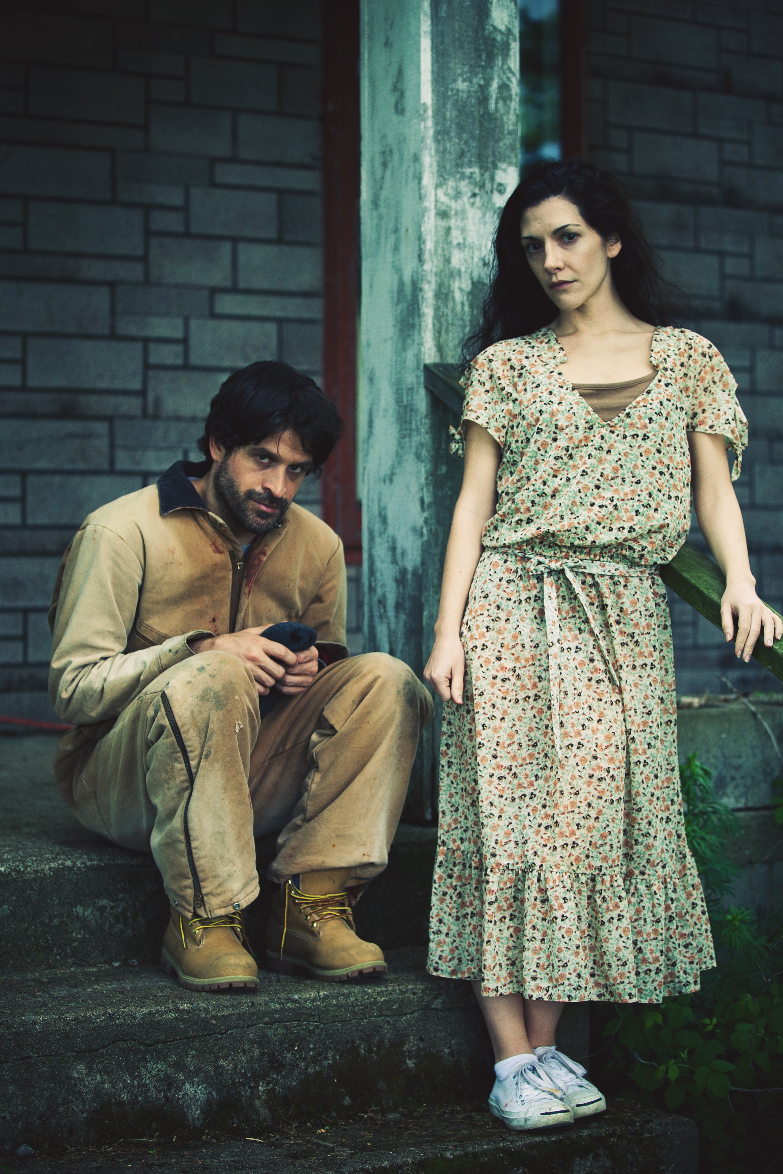 A man with dark hair and a beard sitting on concrete steps, looking at his phone, wearing tan work clothes and boots. A woman with dark hair and a serious expression, standing next to him, wearing a floral dress and white sneakers, leaning against a 