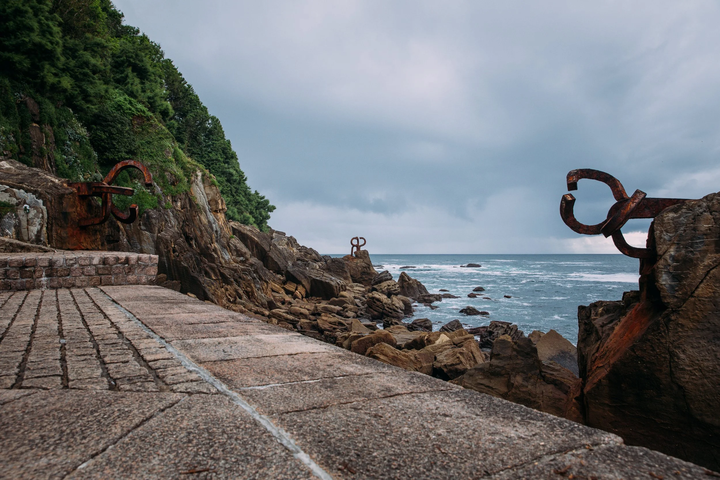 A rocky seaside walkway with rusted metal Sculptures on the rocks and behind a brick-paved path, overlooking the ocean under a cloudy sky.