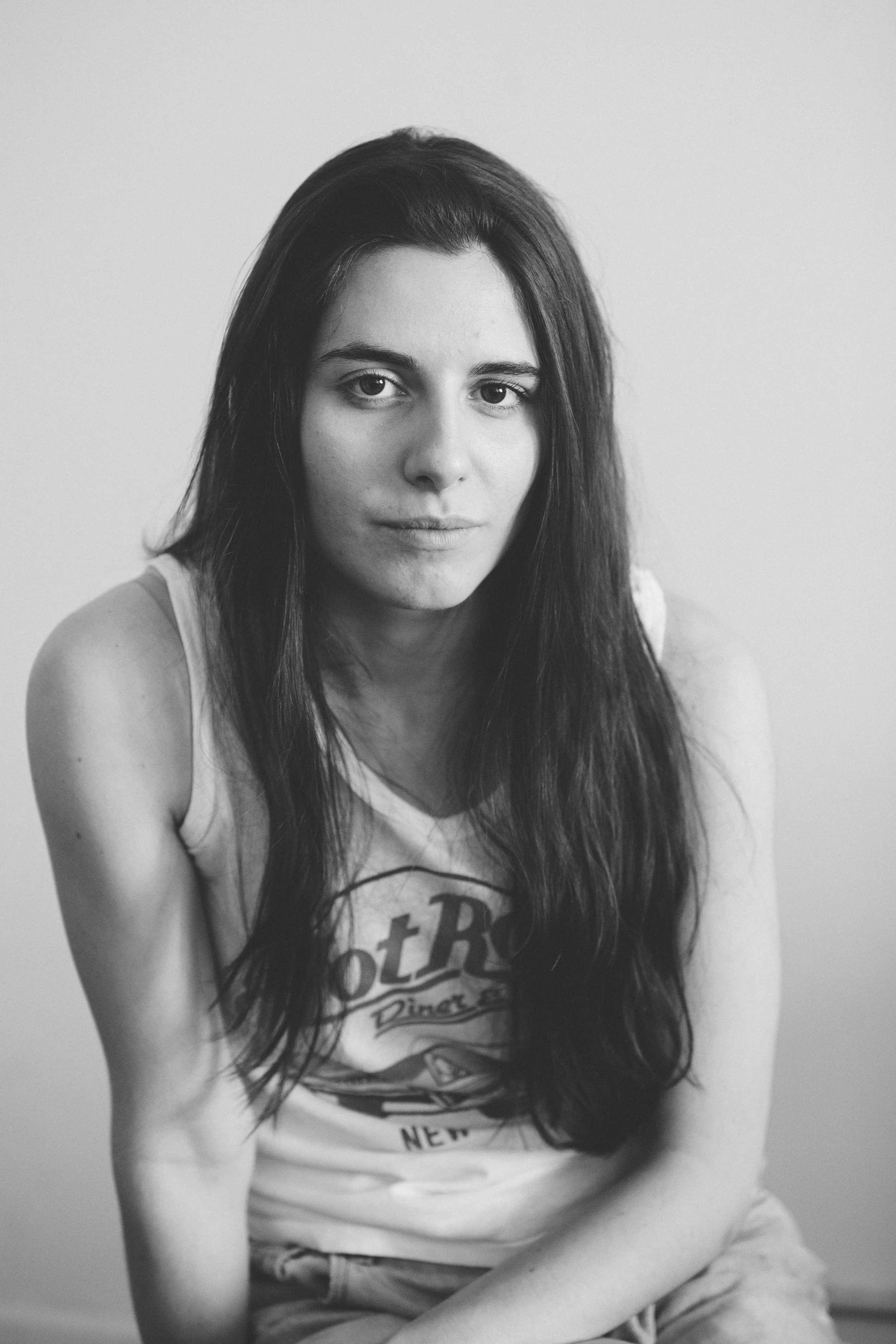 A black and white portrait of a young woman with long dark hair, wearing a sleeveless shirt, looking directly at the camera with a neutral expression.