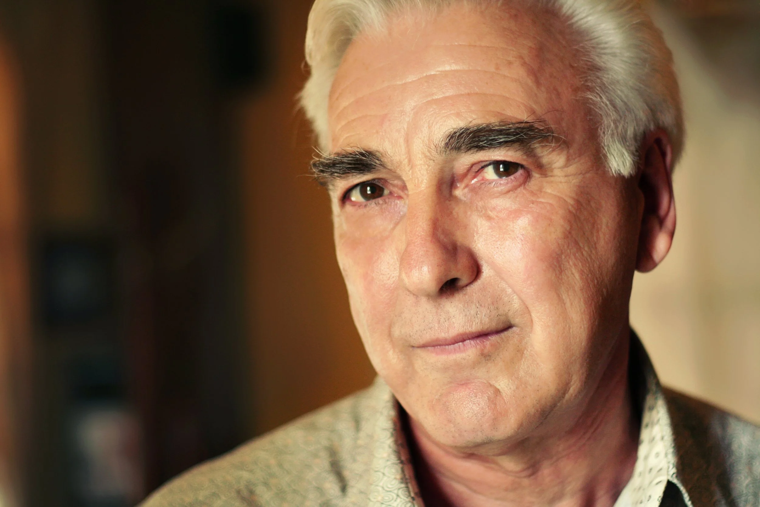 Close-up of an older man with grey hair and dressed in a patterned shirt, looking towards the camera with a subtle smile.