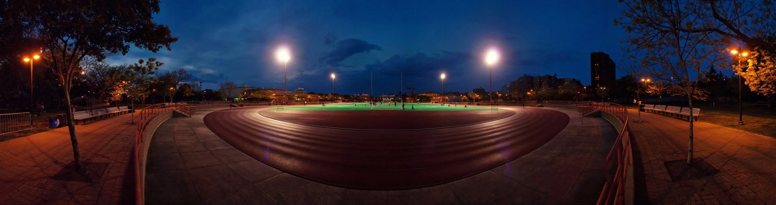 Empty outdoor running track at dusk with bright stadium lights and surrounding trees, city skyline in the background