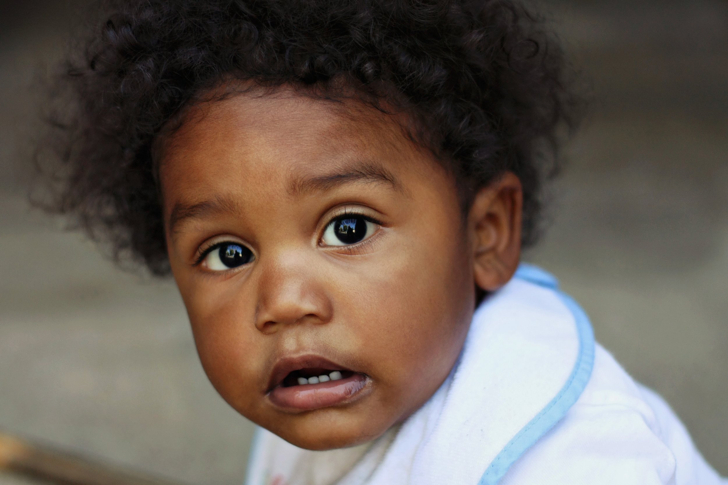 Close-up of a young child's face with dark curly hair, wide eyes, and an expression of surprise or curiosity, wearing a light blue shirt.