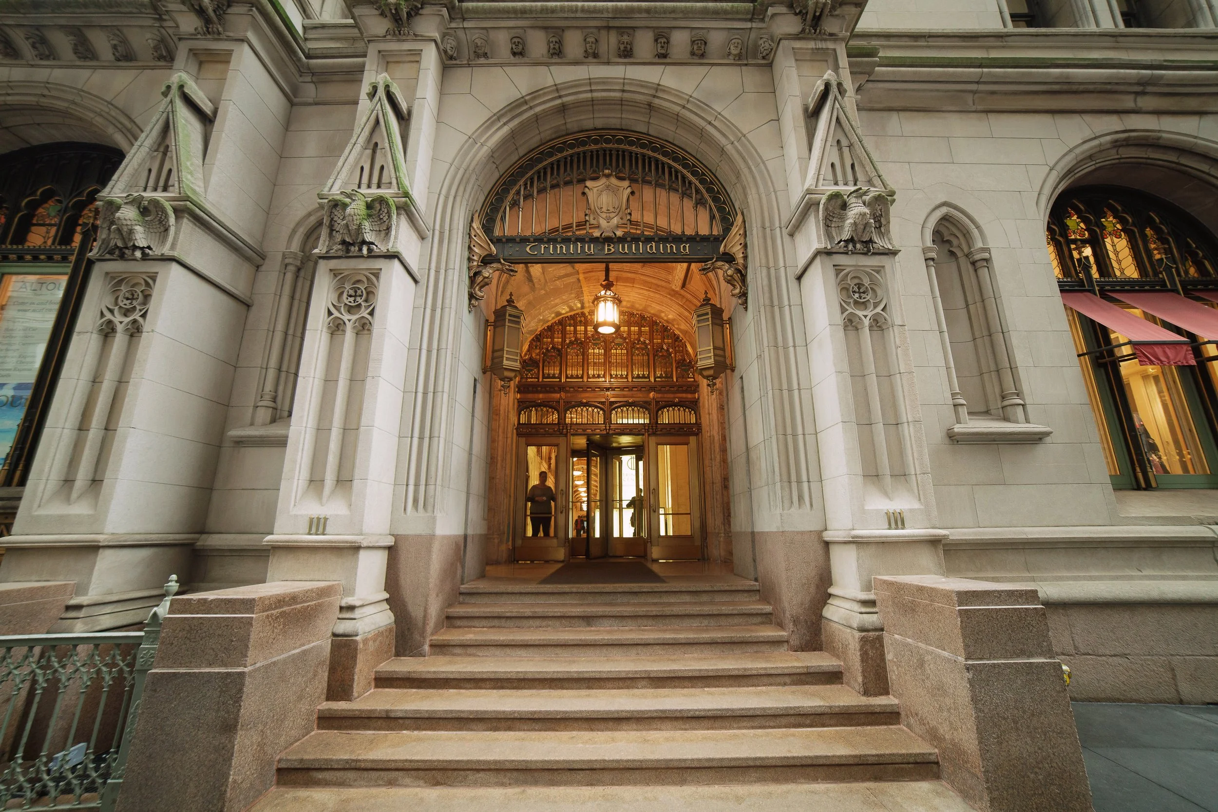 Entrance to the Trinity Building featuring stone architecture, ornate carvings, and glass doors, with steps leading up to the doorway.