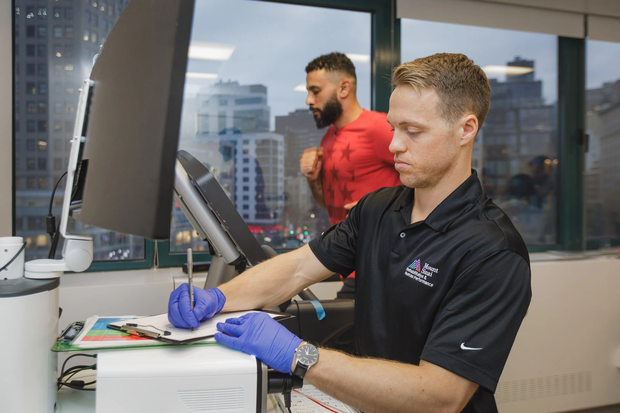 Two men working in a laboratory or medical facility. The man in the foreground is wearing a black polo shirt with a logo that reads "Mount Sinai Rehabilitation & Human Performance" and is wearing blue gloves while working with laboratory equipment. T
